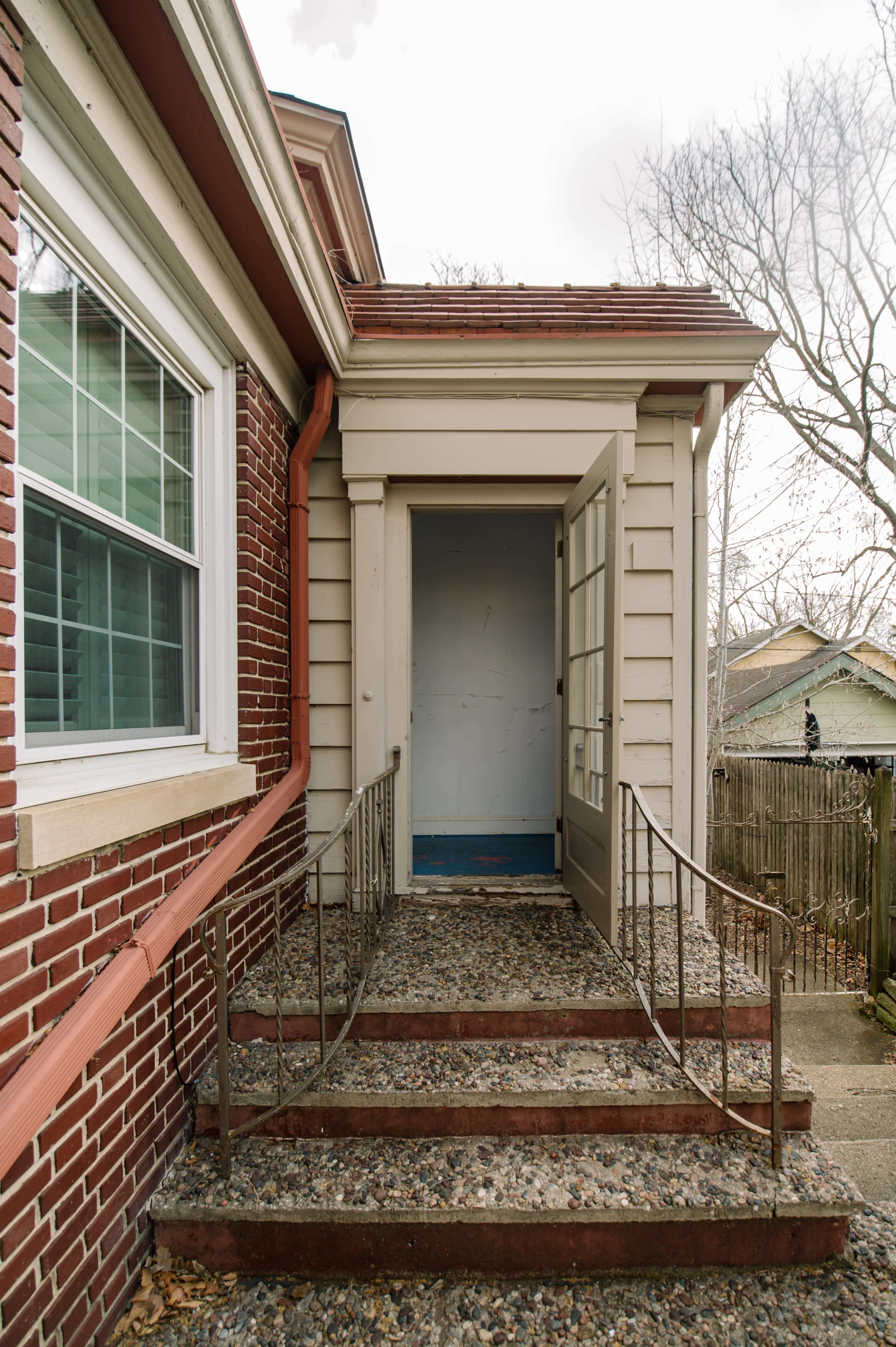 A brick house with a small set of steps leading to an open door, framed by a railing and surrounded by a gravel pathway.
