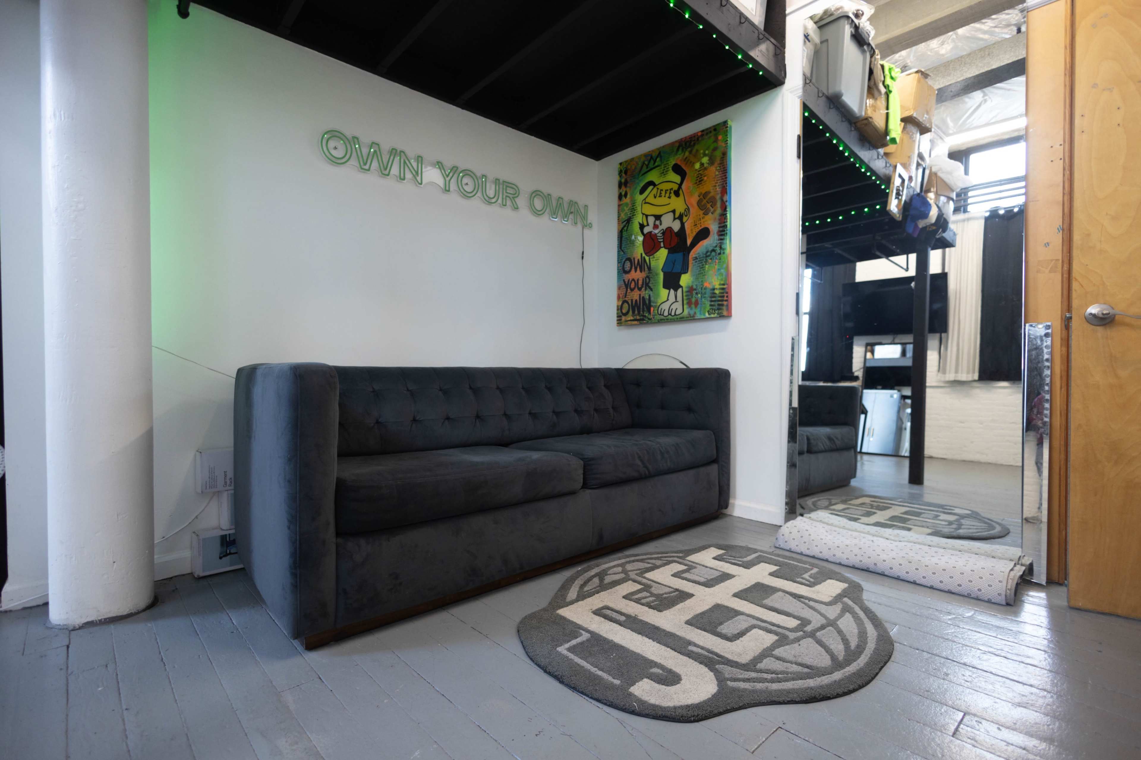 The image shows a modern interior with a gray sofa, a large rug featuring the name "JEFF," and an artwork of a cartoon character on the wall.