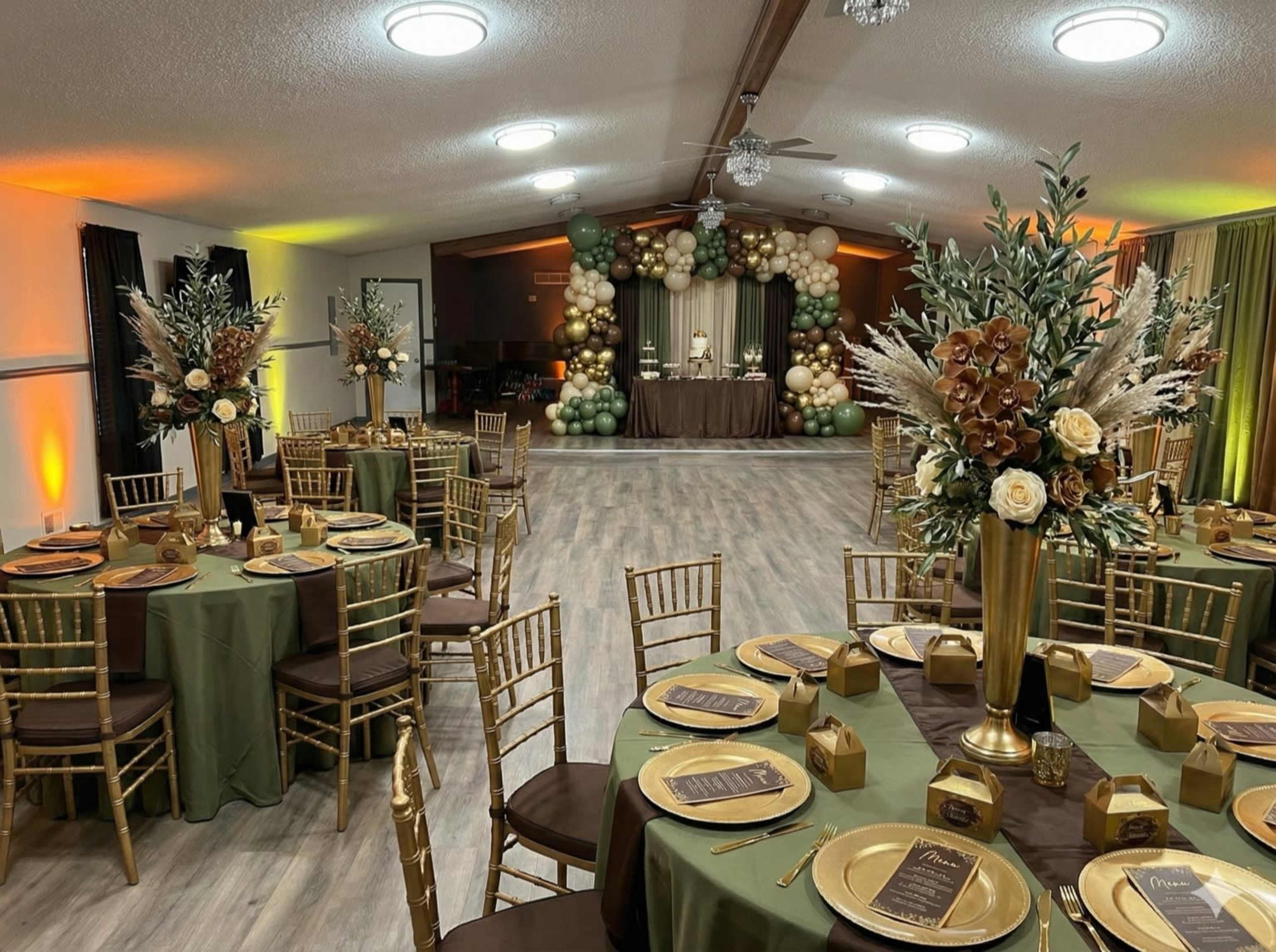 The image shows a decorated indoor event space with elegantly set tables featuring golden chairs, floral centerpieces, and a backdrop of balloons.