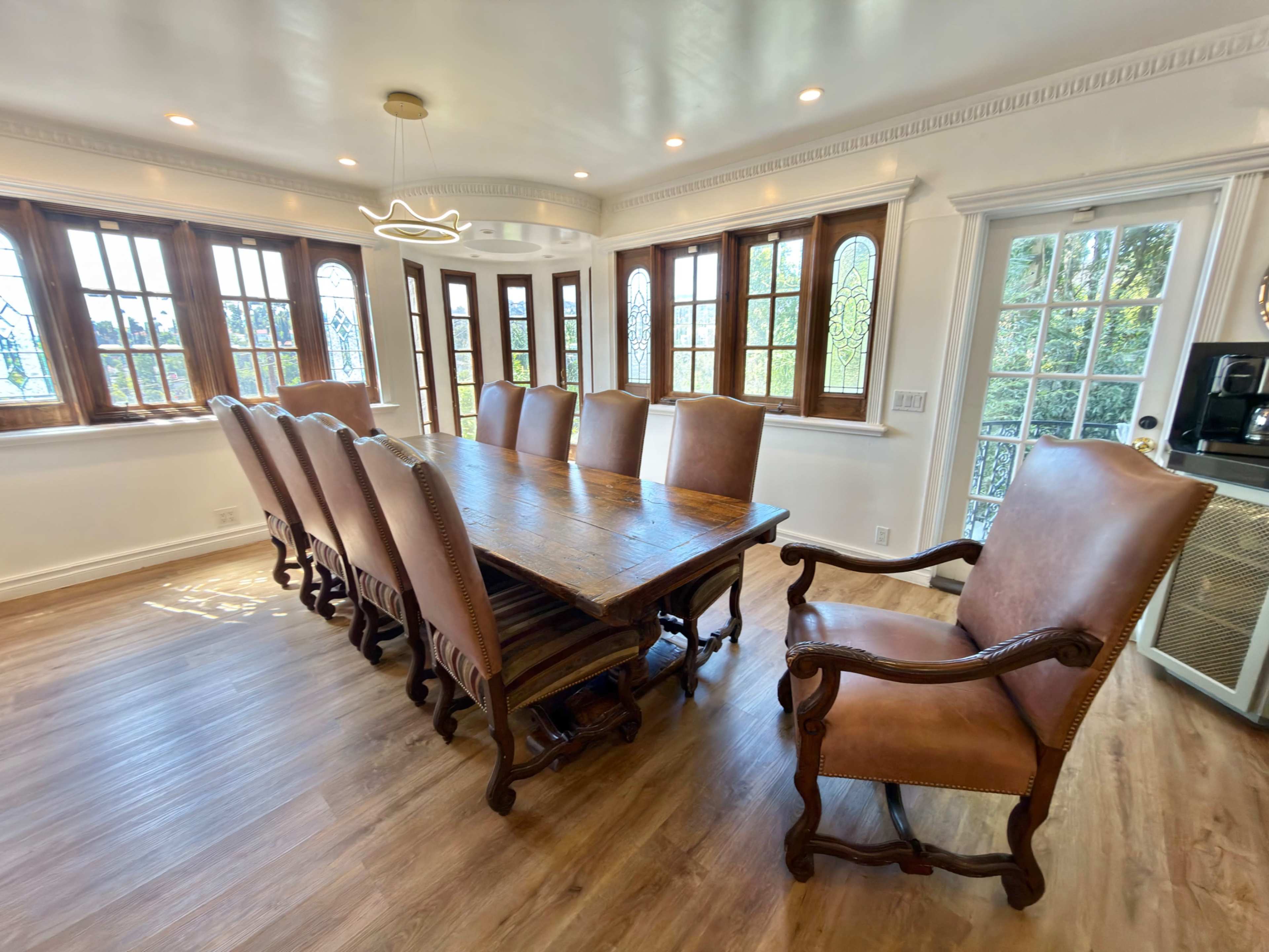 The image shows a spacious dining room with a large wooden table and eight brown leather chairs arranged around it, set against a backdrop of large windows that let in natural light.