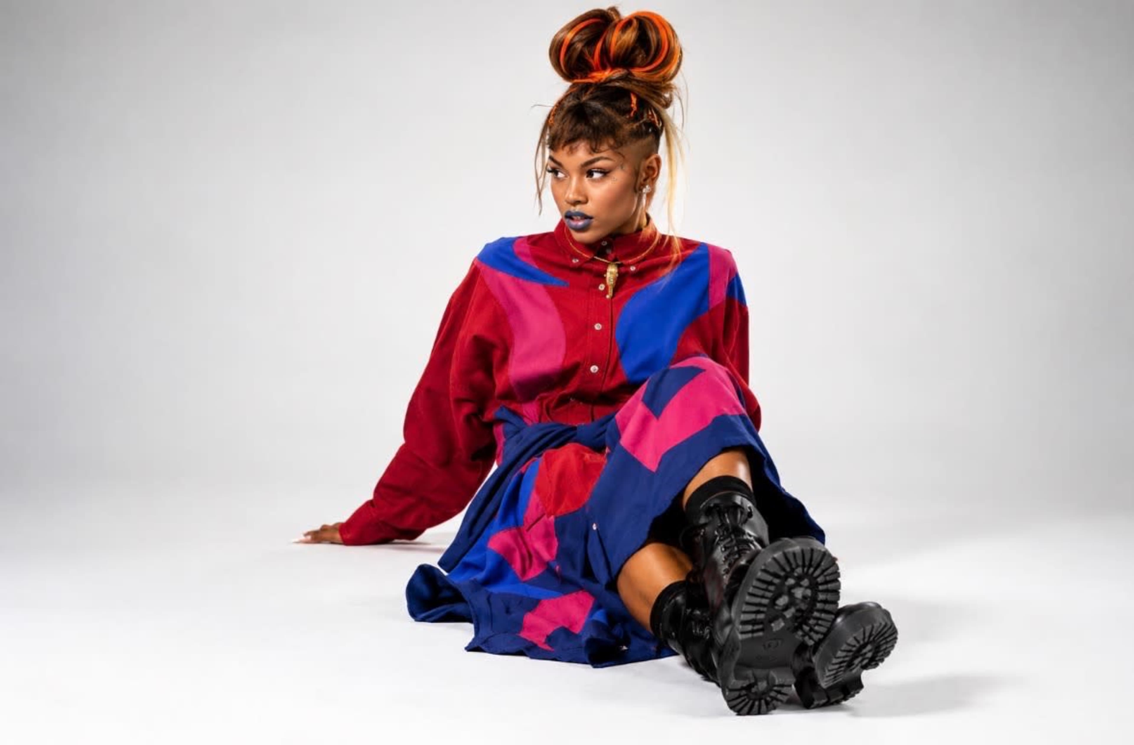 A person sits on the floor, wearing a colorful outfit with a long skirt and combat boots, against a plain gray backdrop.