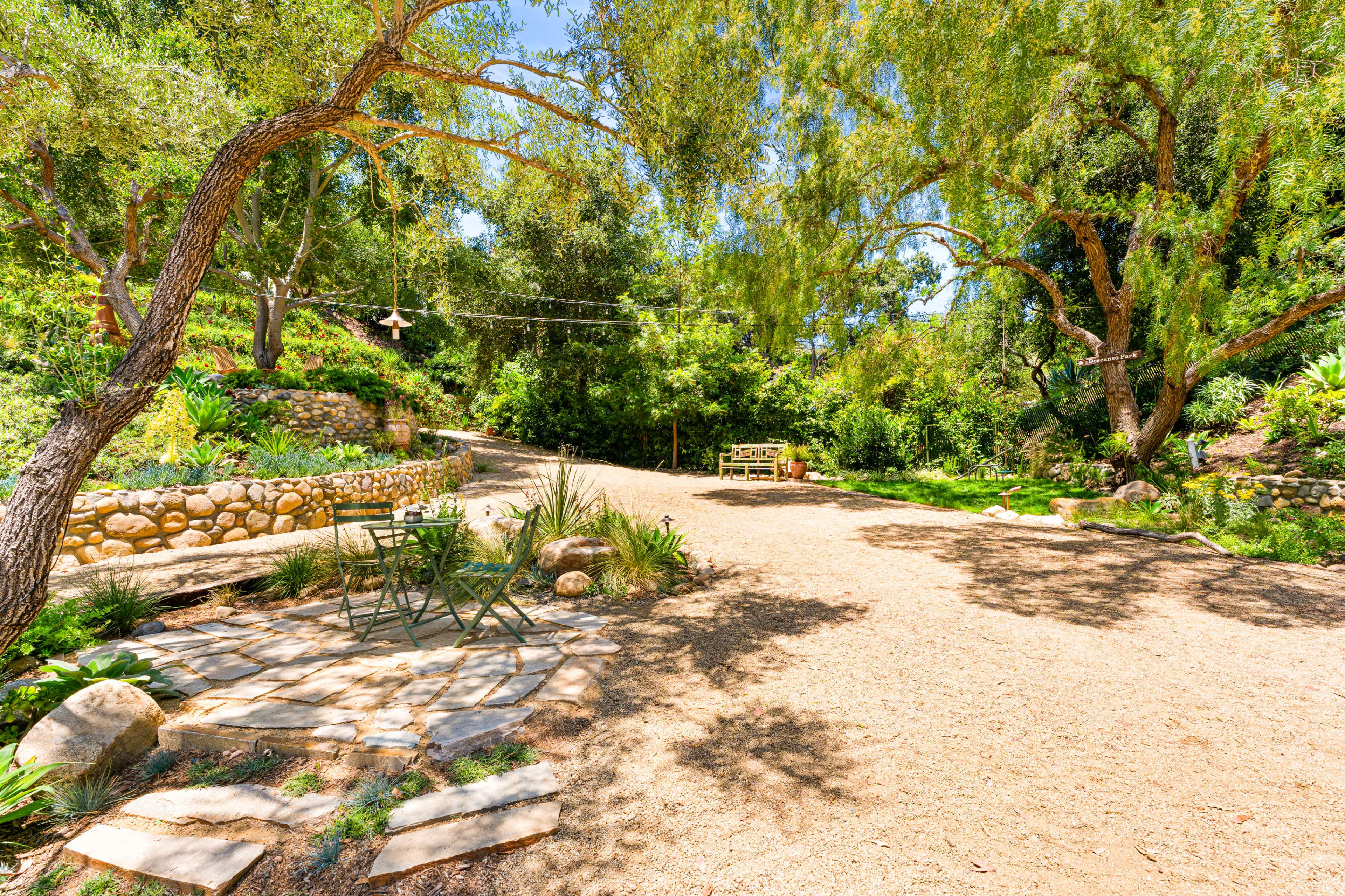 A gravel path winds through a landscaped garden with stone walls, trees, and a bench in the background.