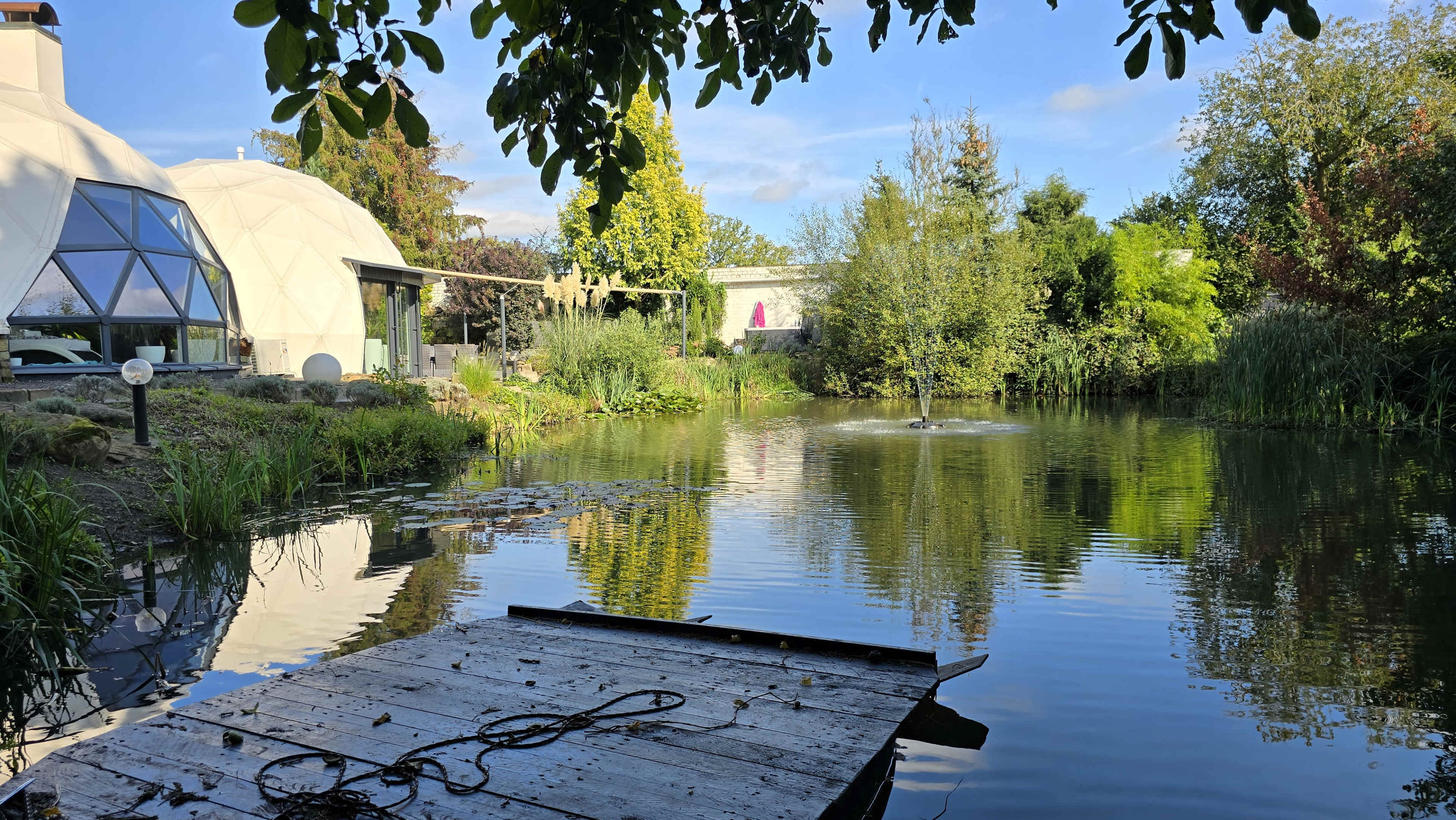 A wooden platform extends over a calm pond surrounded by greenery and a geodesic dome structure in the background.