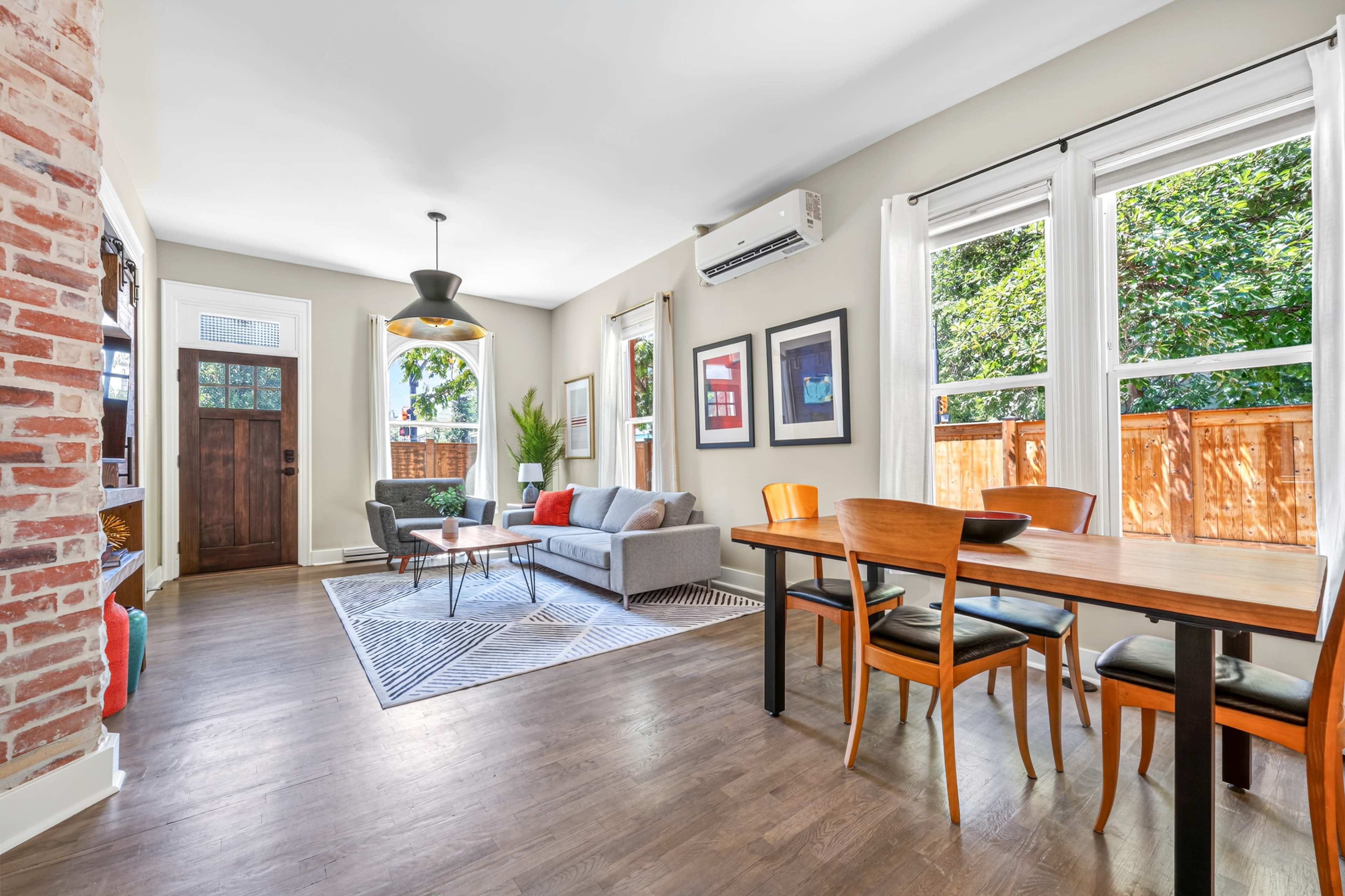 The image shows a bright living room with a gray sofa, a dining table with chairs, and large windows letting in natural light.
