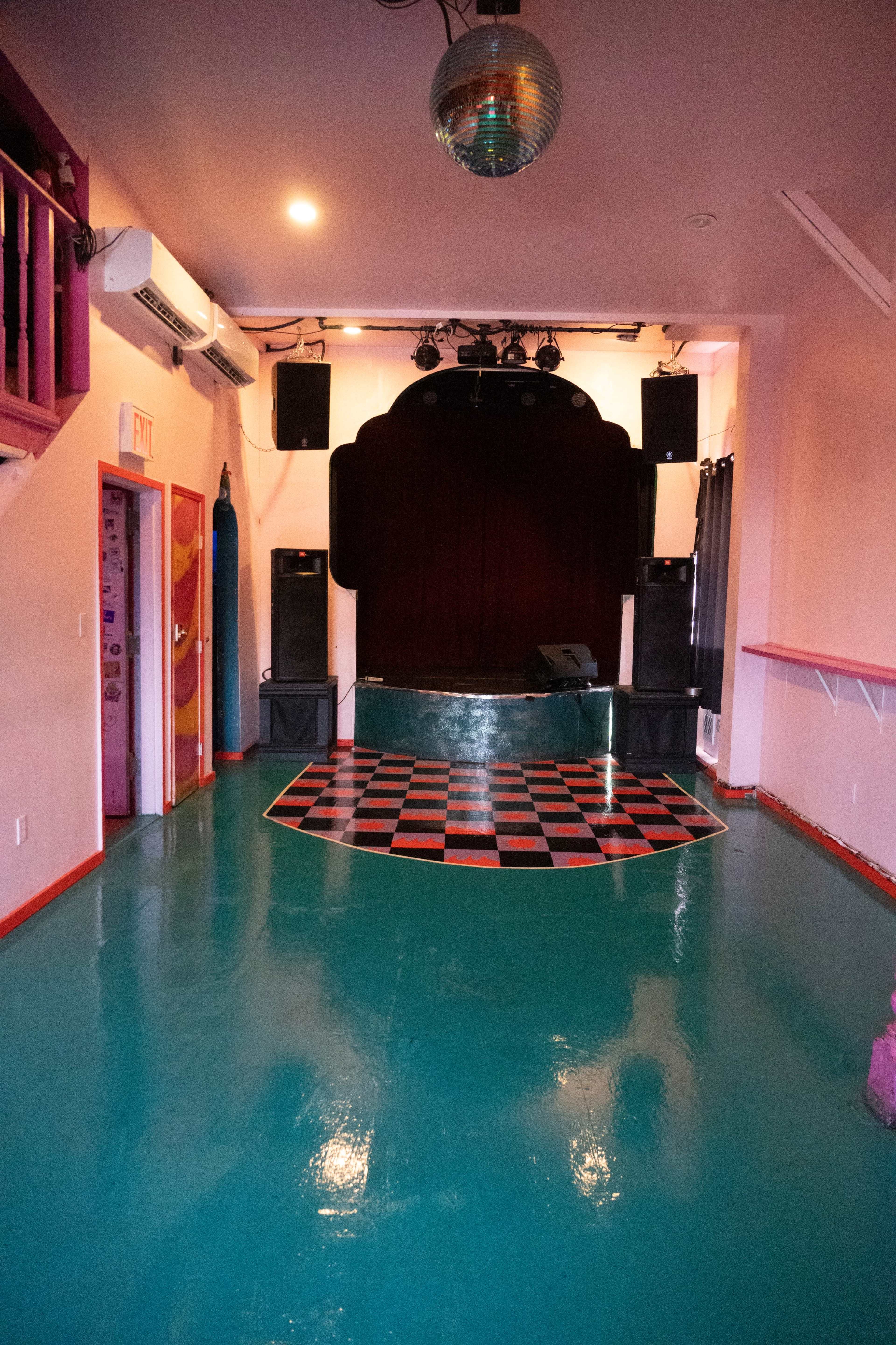 A small performance space with a checkered floor, a stage at the back, and sound equipment setup on either side.