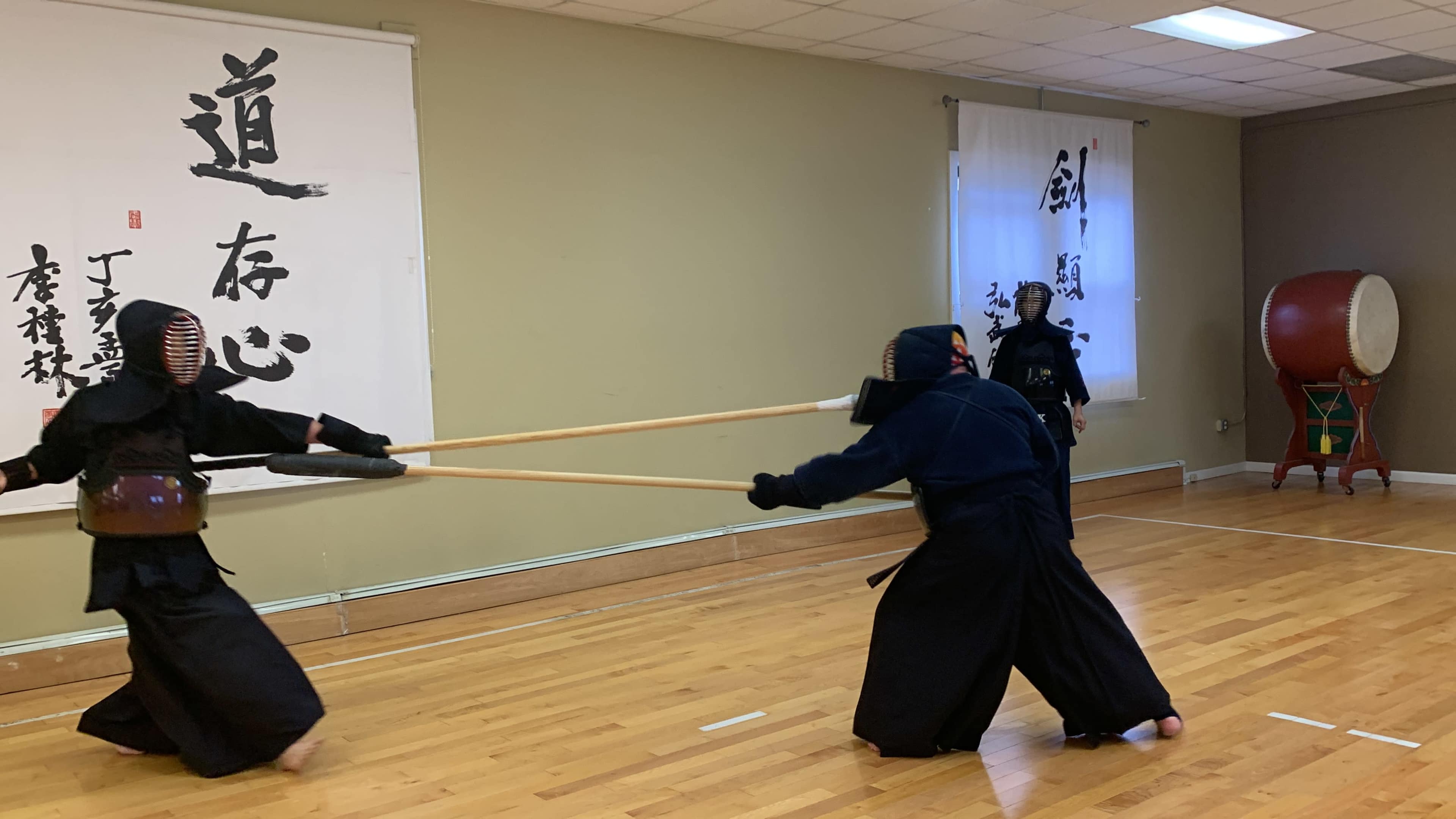 Three individuals in dark traditional clothing practice techniques with long wooden staffs in a spacious dojo.