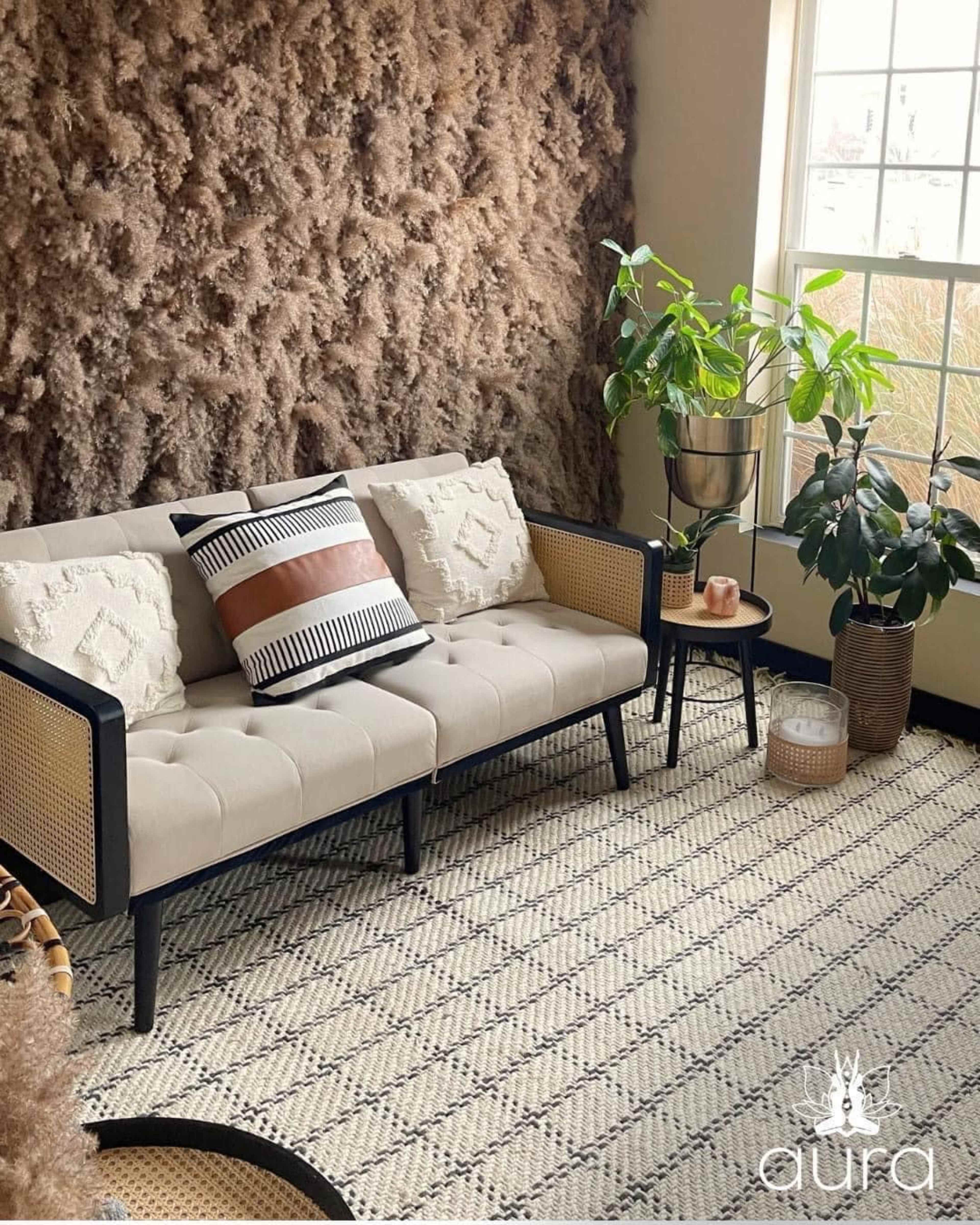 A modern living room features a beige sofa with a patterned pillow, a small side table, and potted plants beside a textured wall and large window.