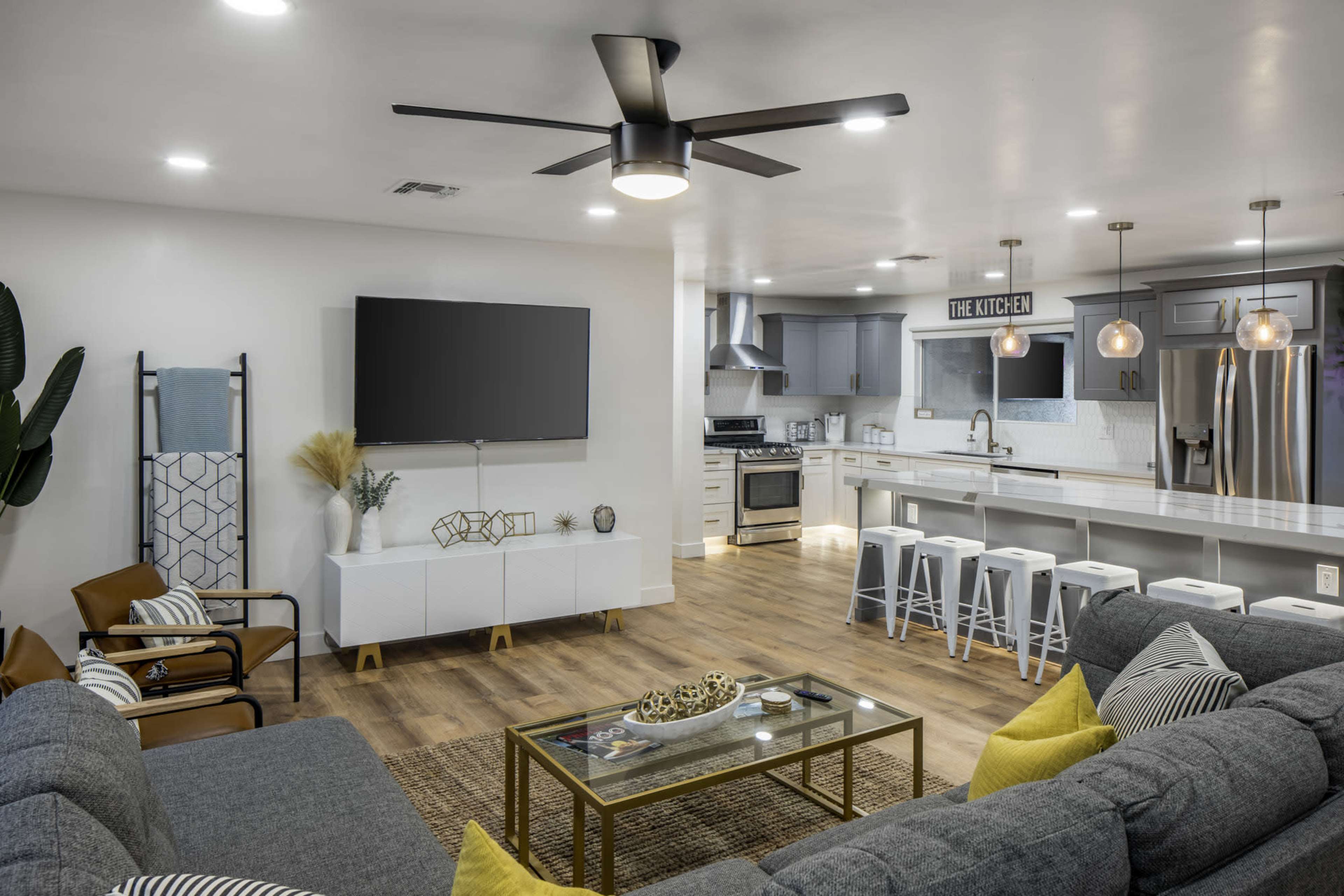 A modern living room and kitchen area with a ceiling fan, large TV, comfortable seating, and a sleek kitchen island with bar stools.