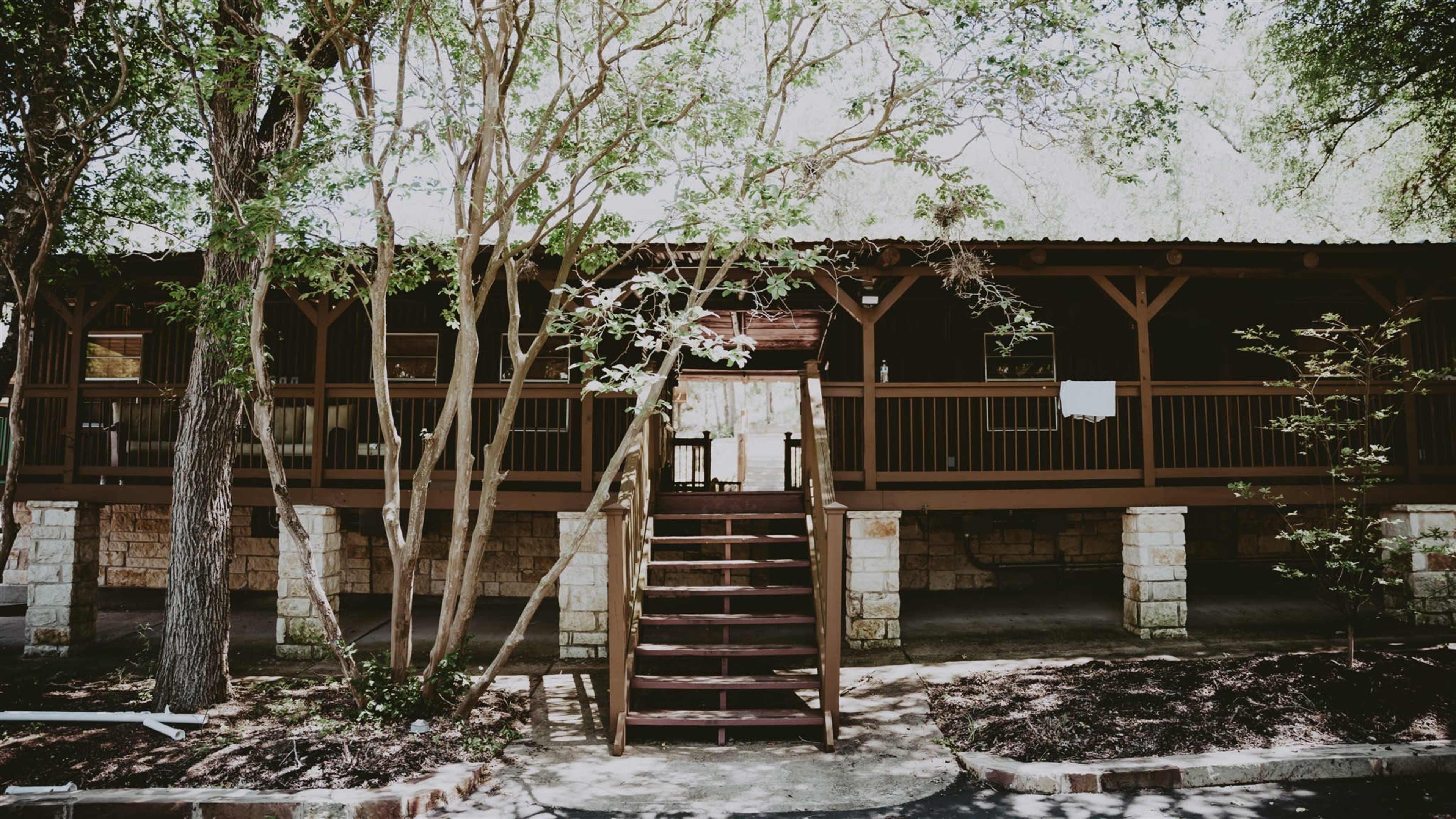 A wooden cabin with a staircase leads up to a porch supported by stone columns, surrounded by trees.