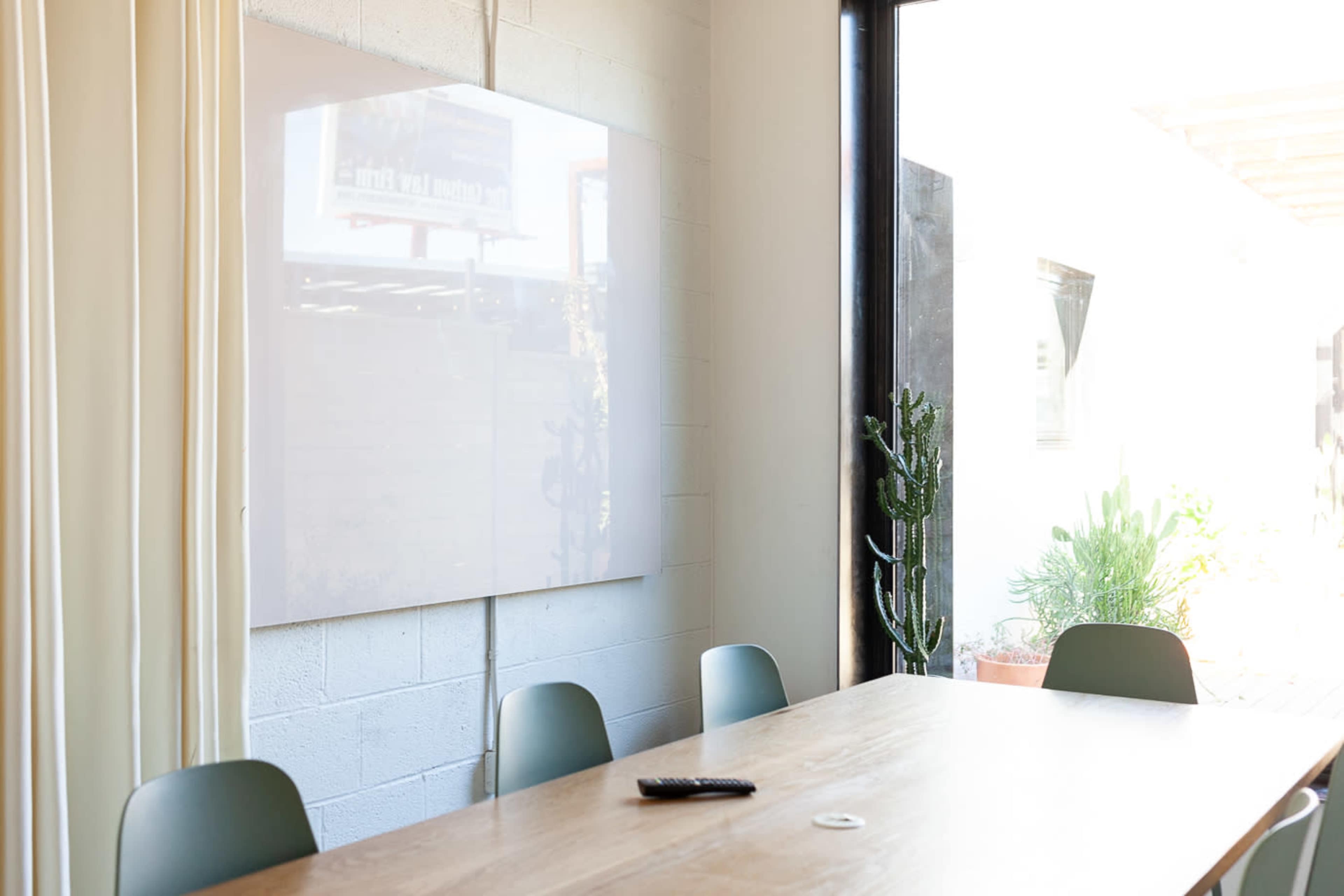 A conference room features a wooden table, chairs, and a large white display on the wall, with plants visible through a large window.