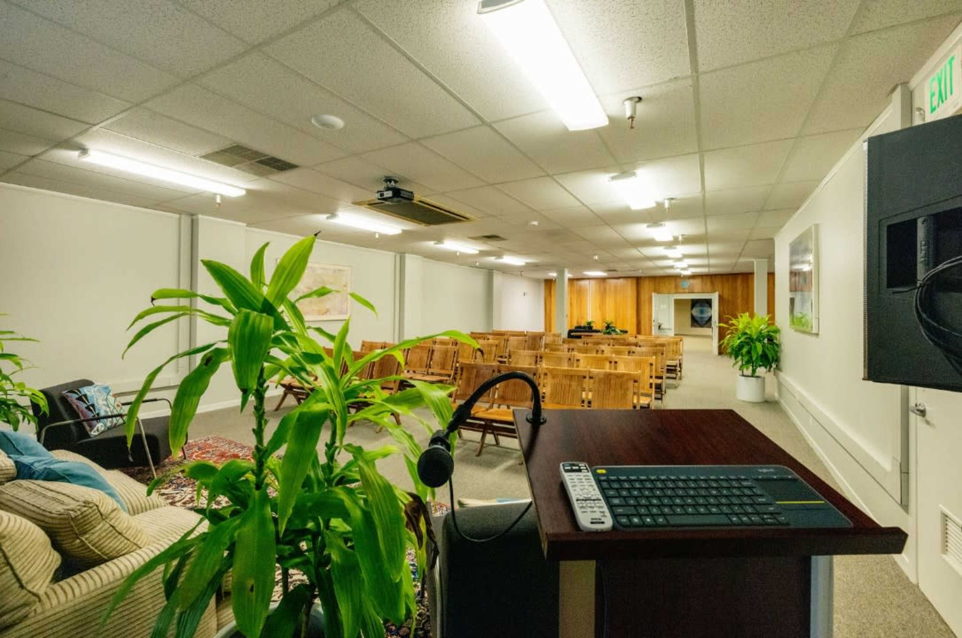 A spacious meeting room with rows of wooden chairs, a control panel in the foreground, and plants scattered around the space.