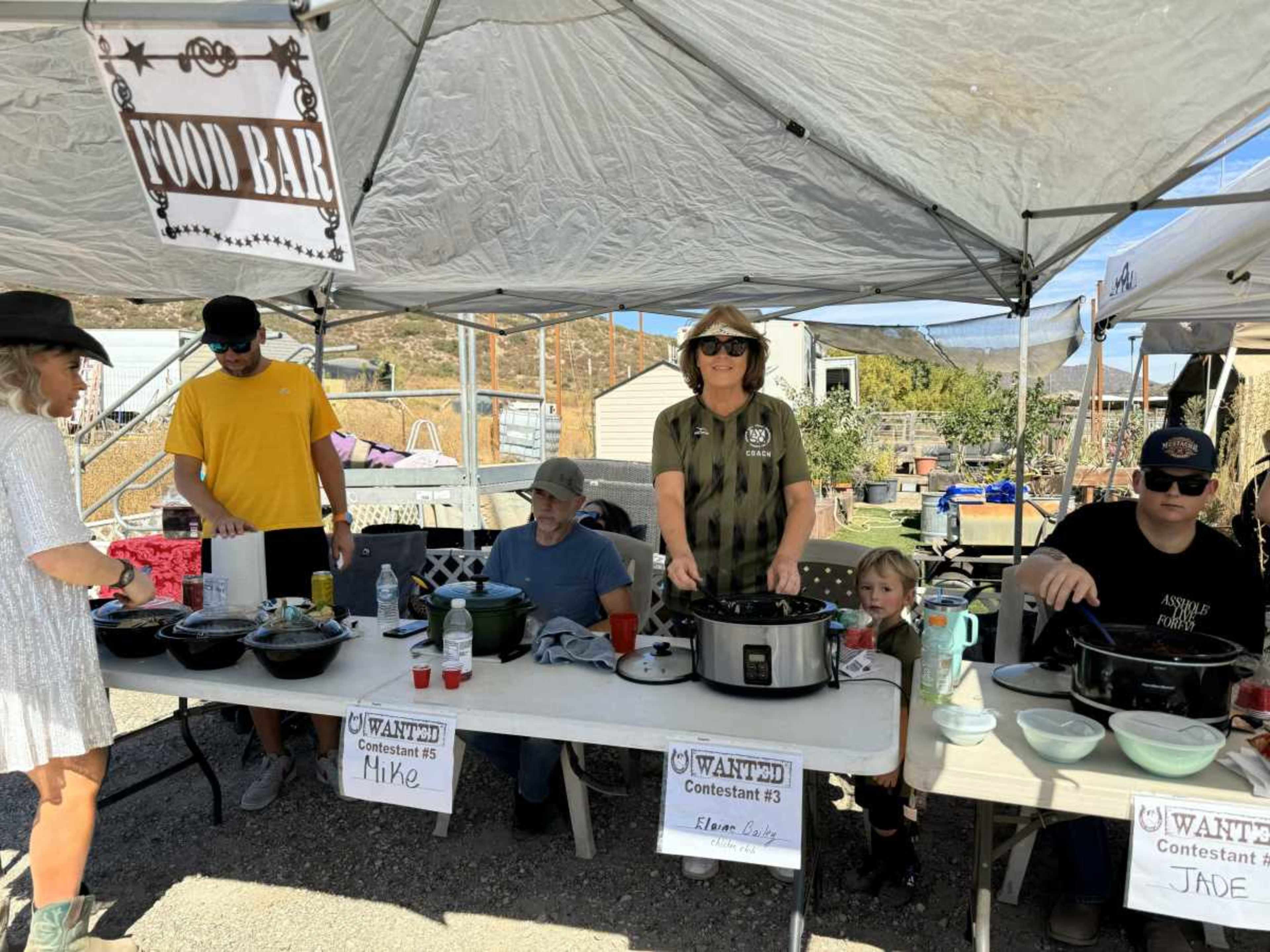 The image shows a food bar at a community event, featuring several contestants behind tables with pots, labels, and a variety of dishes.