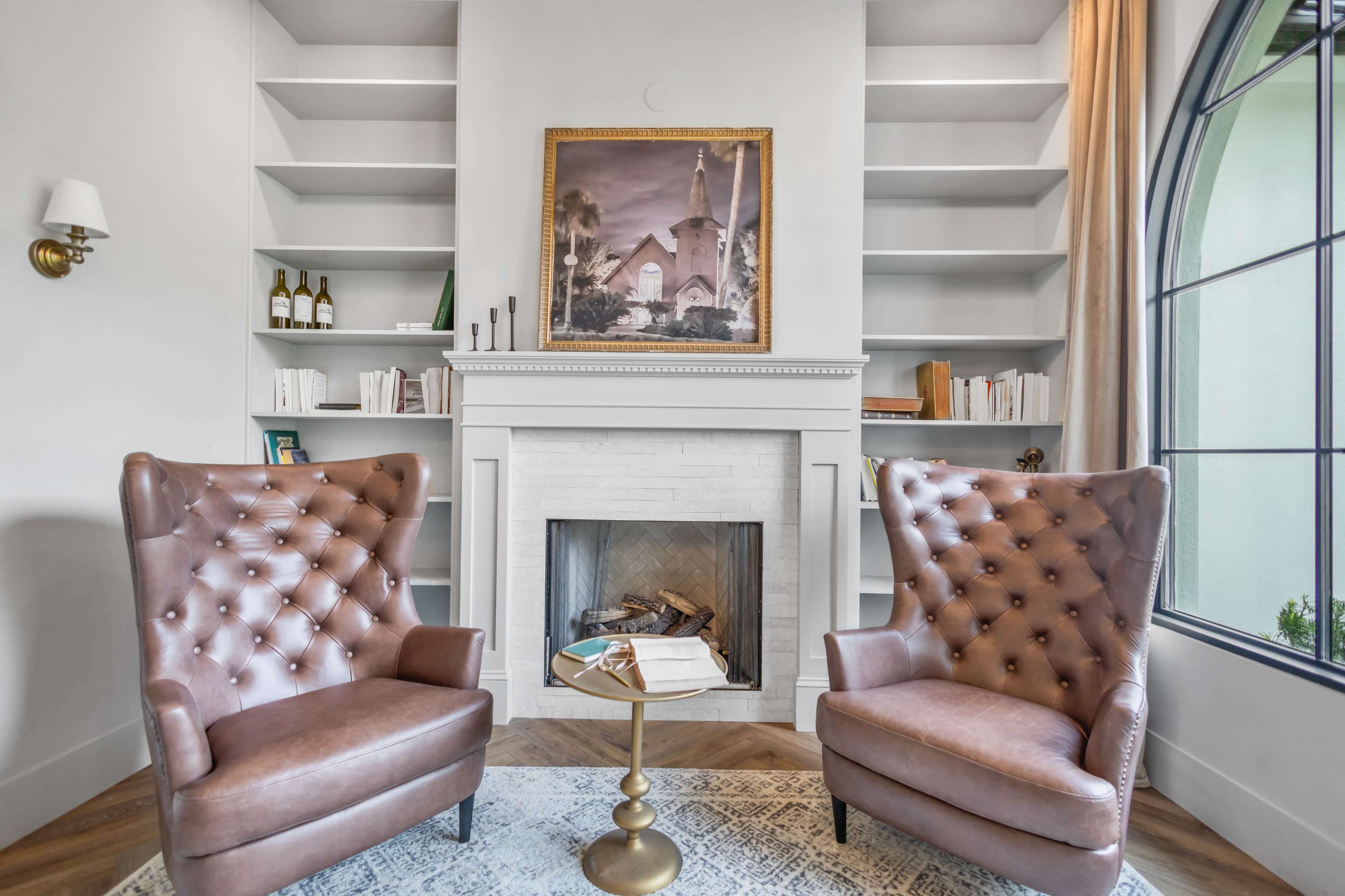 A cozy living space with two tufted brown armchairs facing a small round table, a fireplace surrounded by shelves, and a large window.