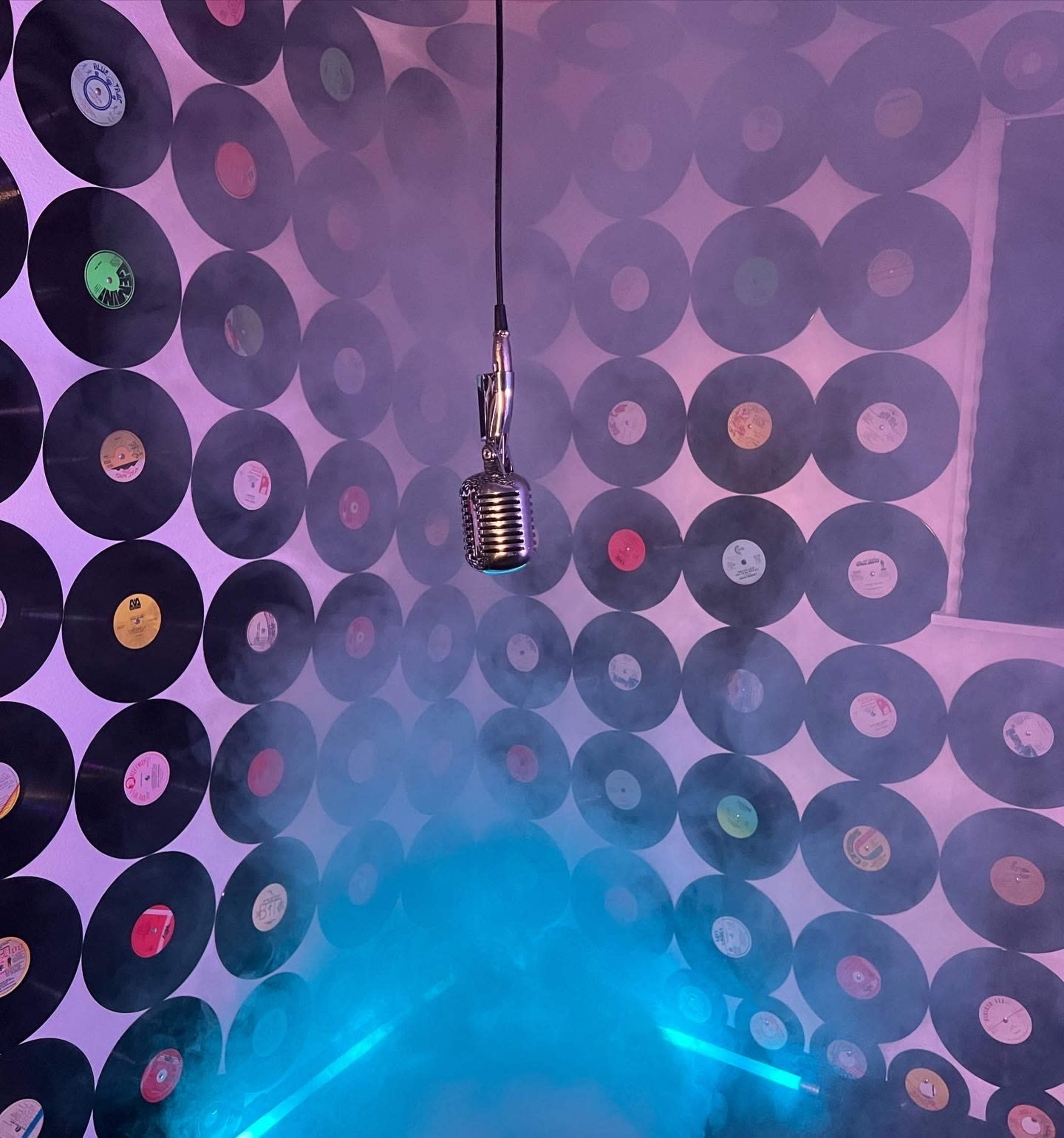 A vintage microphone hangs from the ceiling in a room lined with vinyl records, shrouded in fog and illuminated by blue lighting.