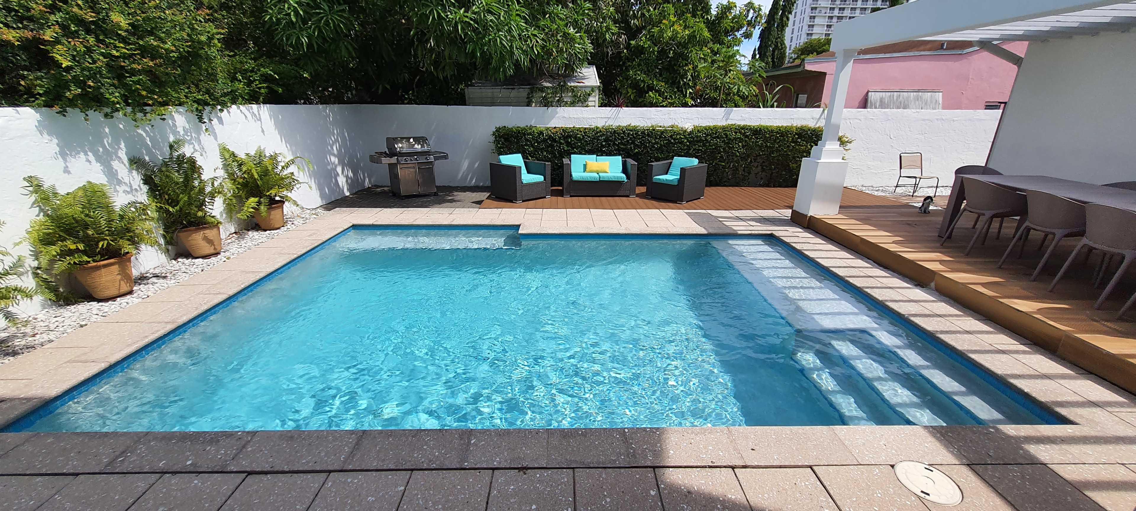 The image shows a rectangular swimming pool surrounded by potted plants, outdoor furniture, and a dining area.