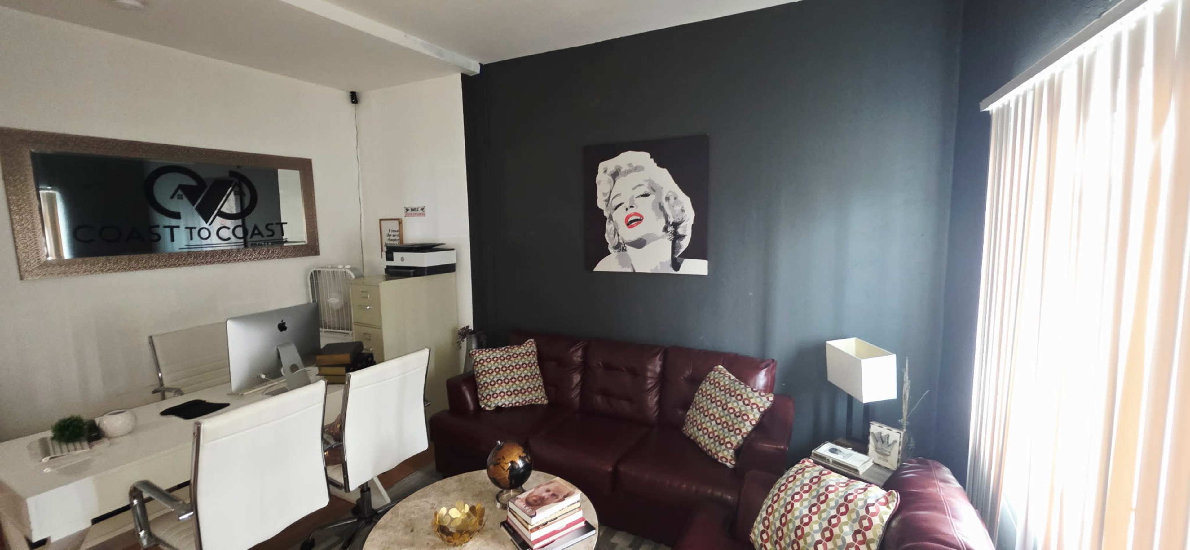A modern living room features a maroon leather sofa, a round coffee table with books, and a large black-and-white artwork of a woman on a gray wall.