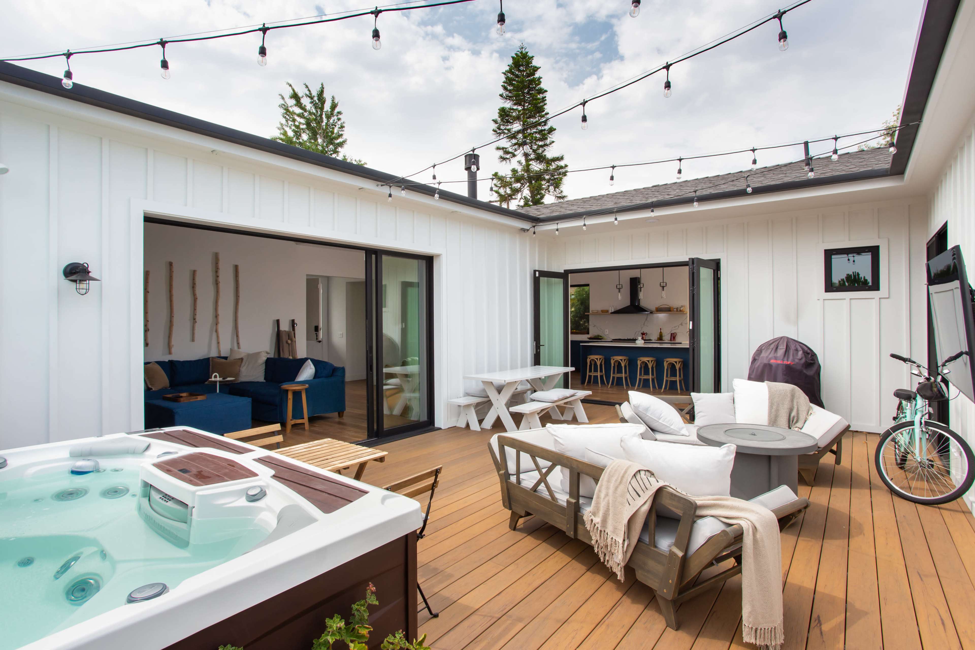 The image shows a modern outdoor patio featuring a hot tub, seating area, and a dining table, all surrounded by wooden decking and framed by white walls and string lights overhead.