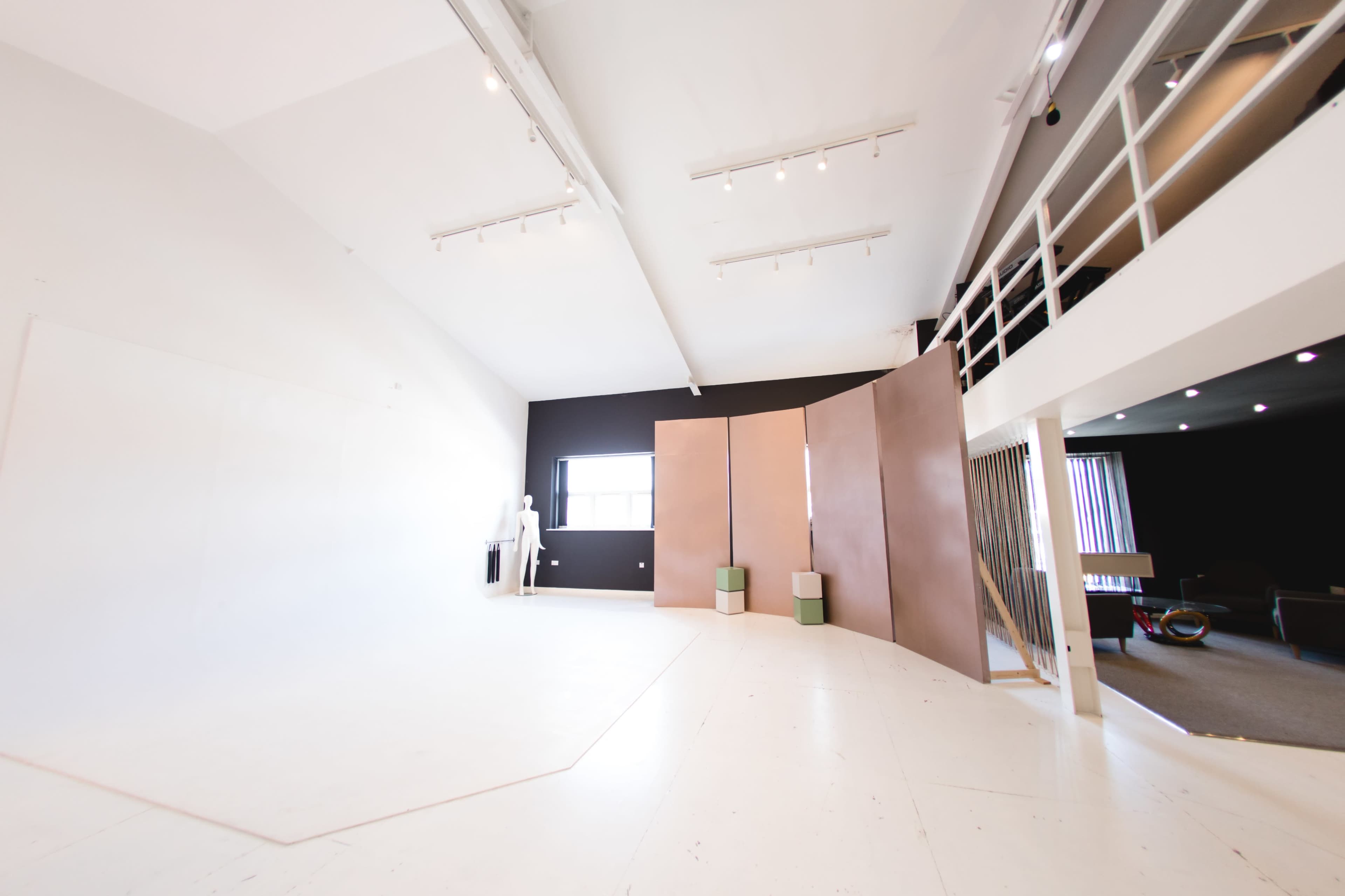A spacious, minimalistic studio with a large white backdrop, light wood flooring, and a dark wall, along with dividing panels and furniture in the background.
