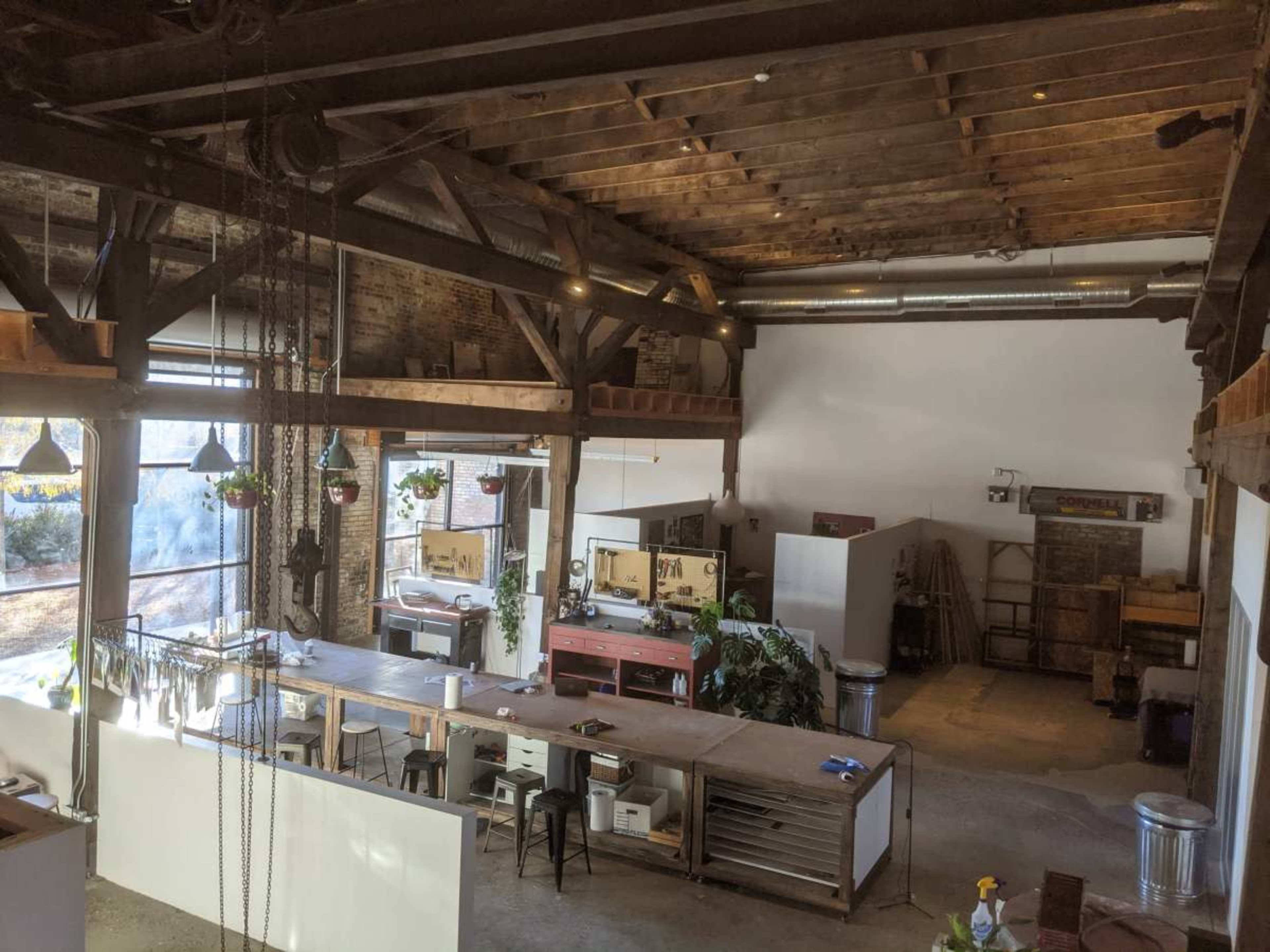 The image shows an open, spacious industrial-style workspace with wooden beams, a large communal table, and various plants hanging from the ceiling.