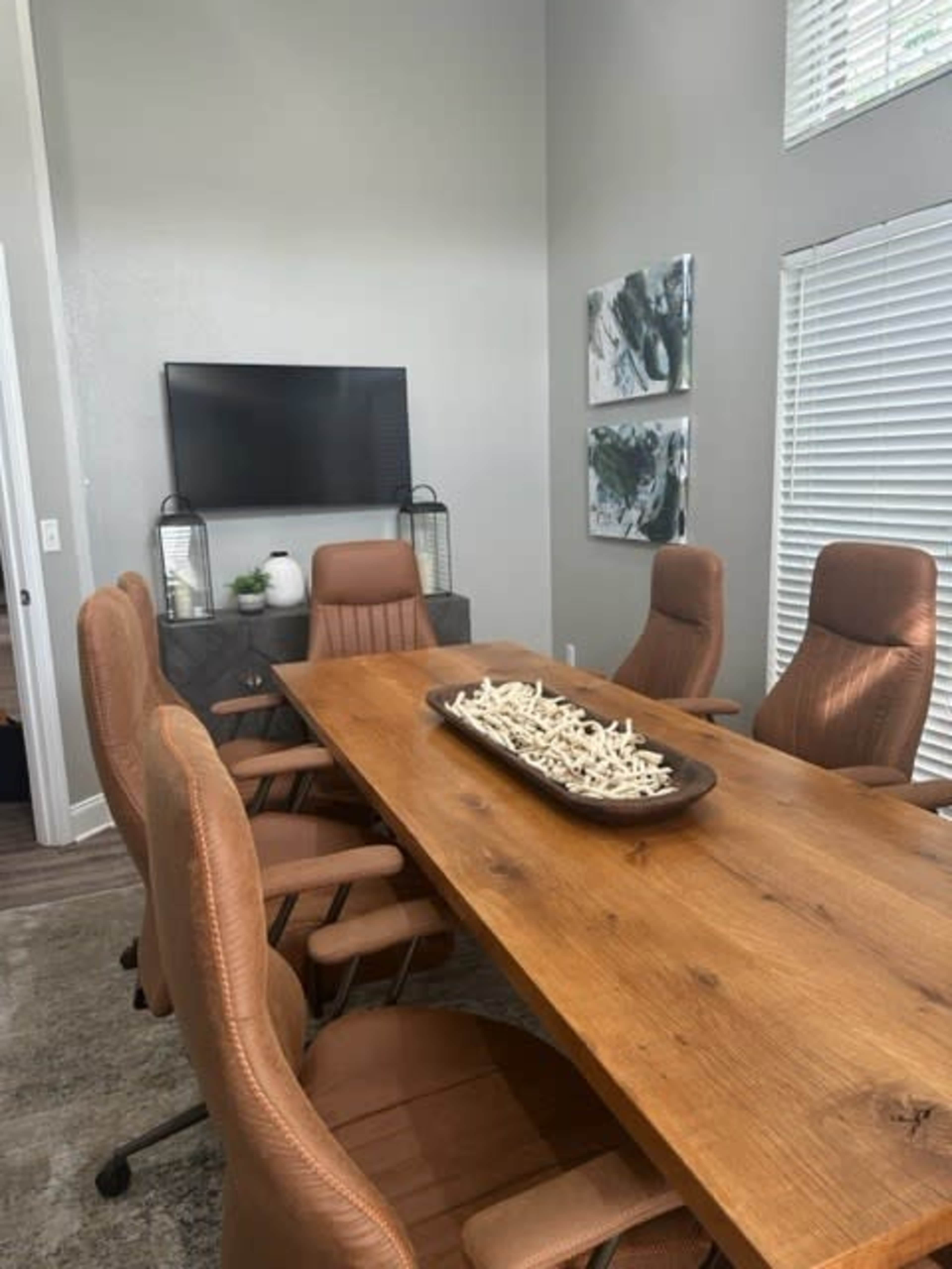 A conference room features a large wooden table surrounded by brown upholstered chairs, with a television mounted on the wall and decorative artwork nearby.