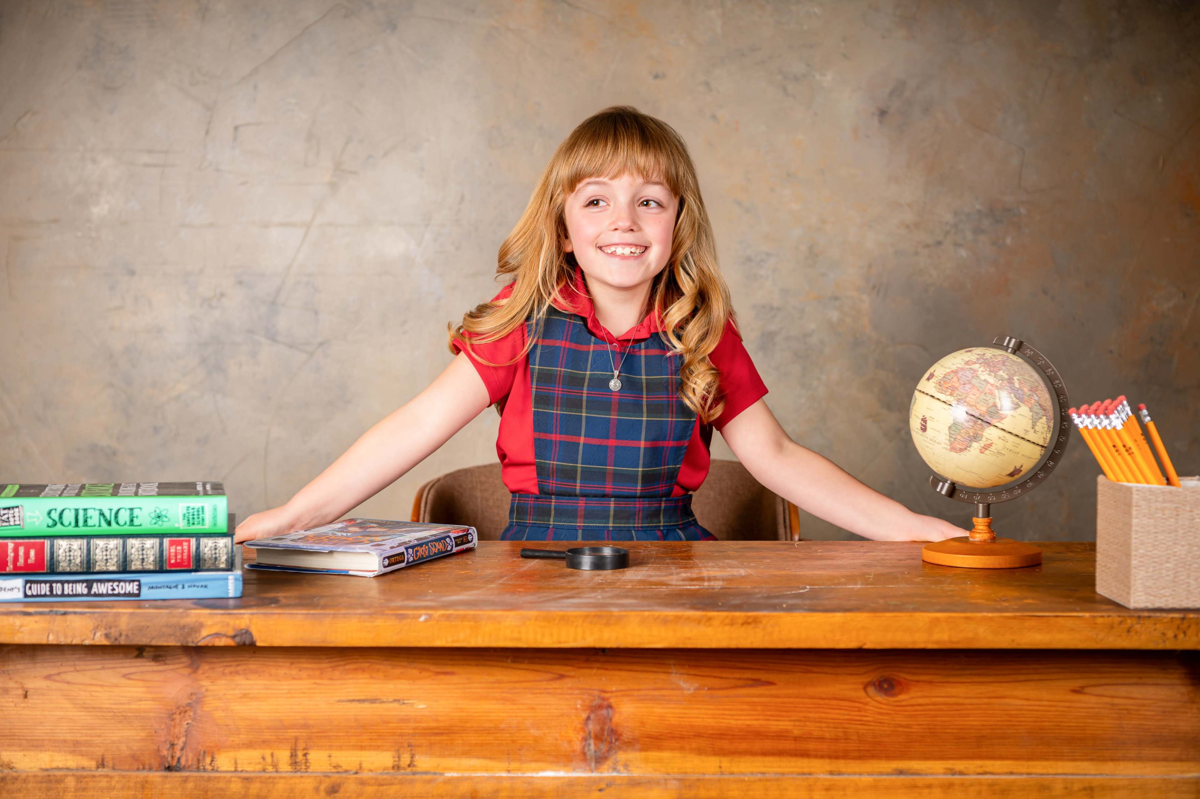 A young girl in a red shirt and plaid dress sits at a wooden desk with books, a globe, and pencils.