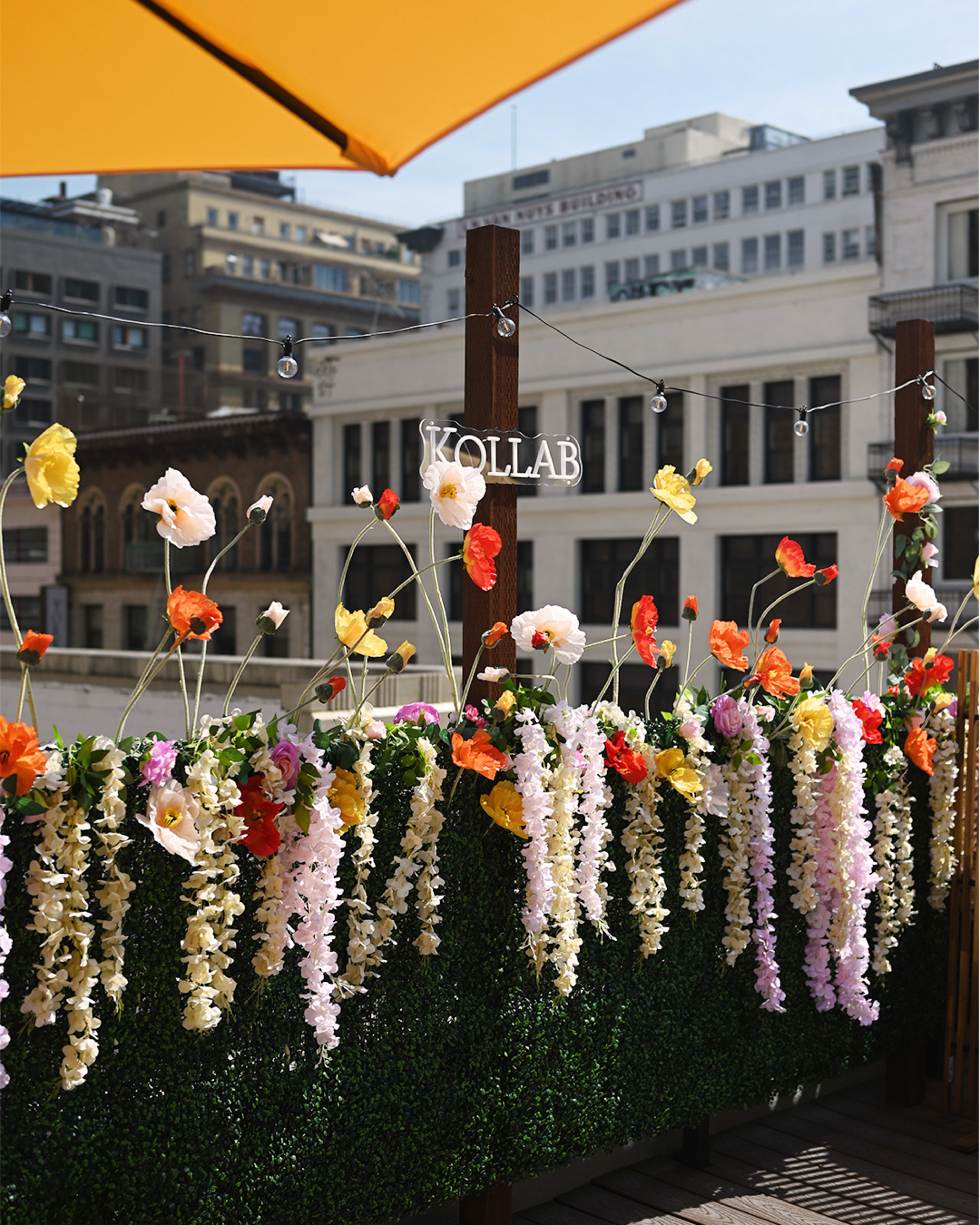The image shows a decorated outdoor space with vibrant, artificial flowers hanging down from a wooden railing, overlooking a cityscape.