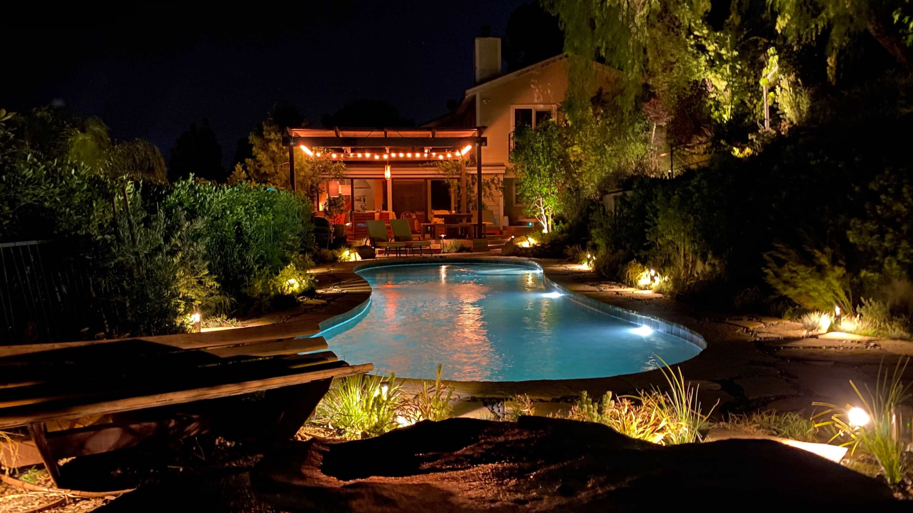 A illuminated outdoor pool is surrounded by lush greenery and a cozy seating area under a covered patio at night.