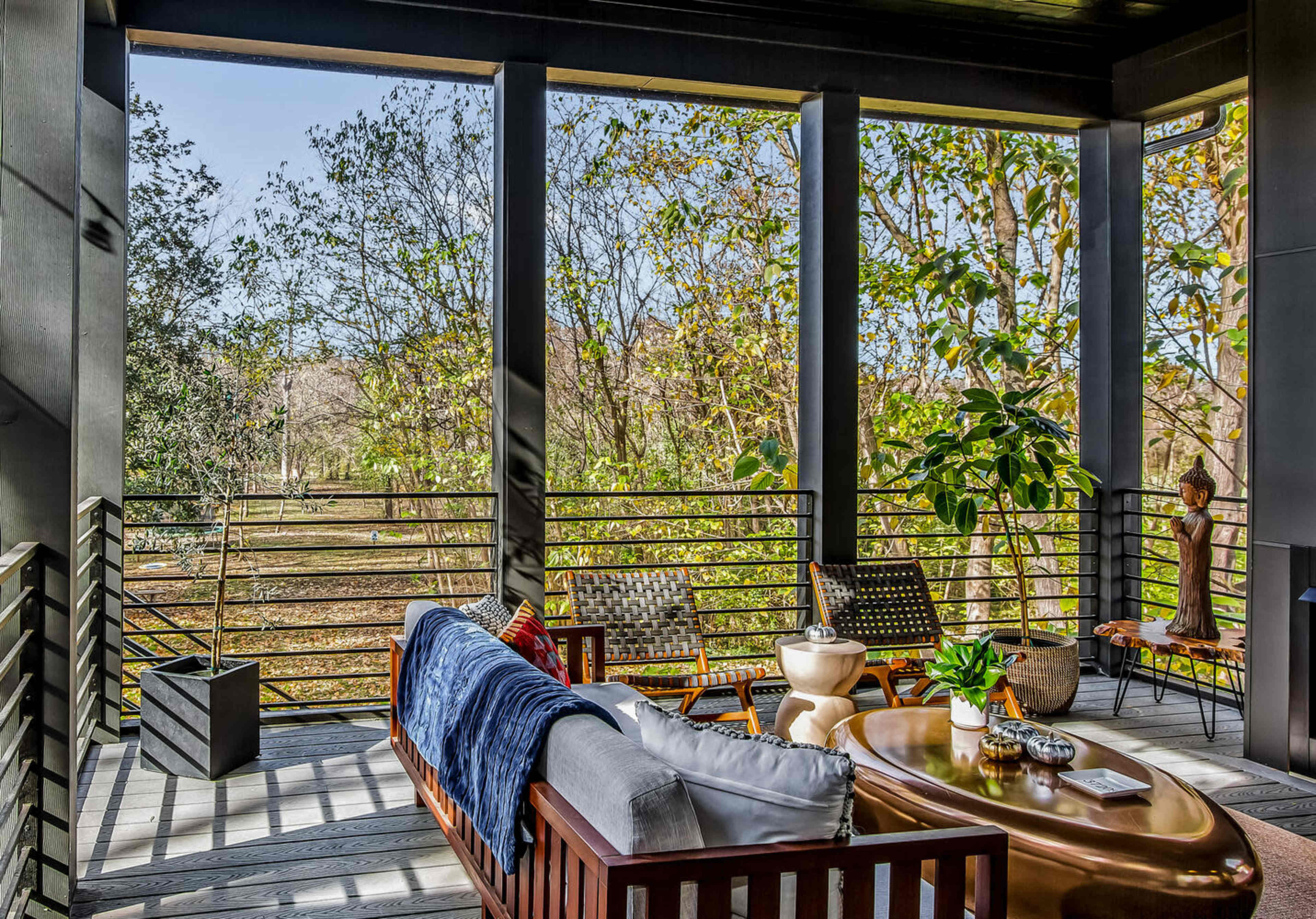 The image shows a modern porch with wooden seating, a round coffee table, and a view of trees and greenery through open railings.