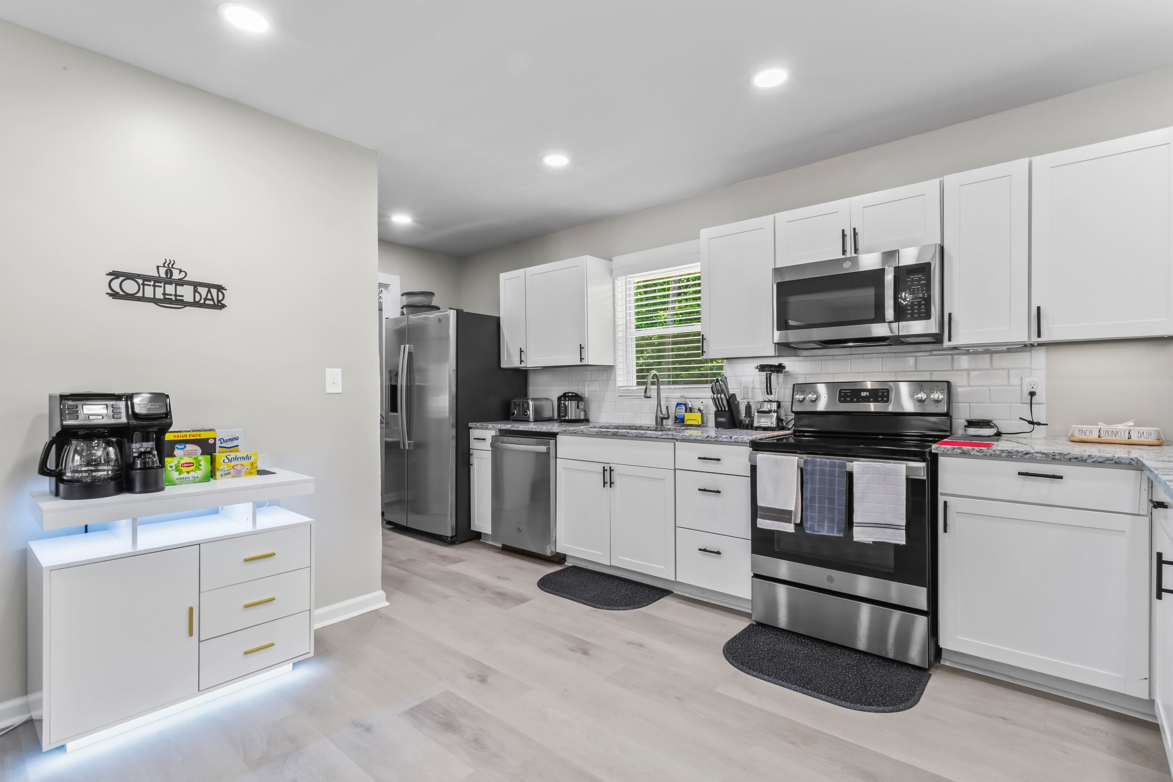 The image shows a modern kitchen featuring white cabinets, stainless steel appliances, and a central island with a coffee maker and snacks.
