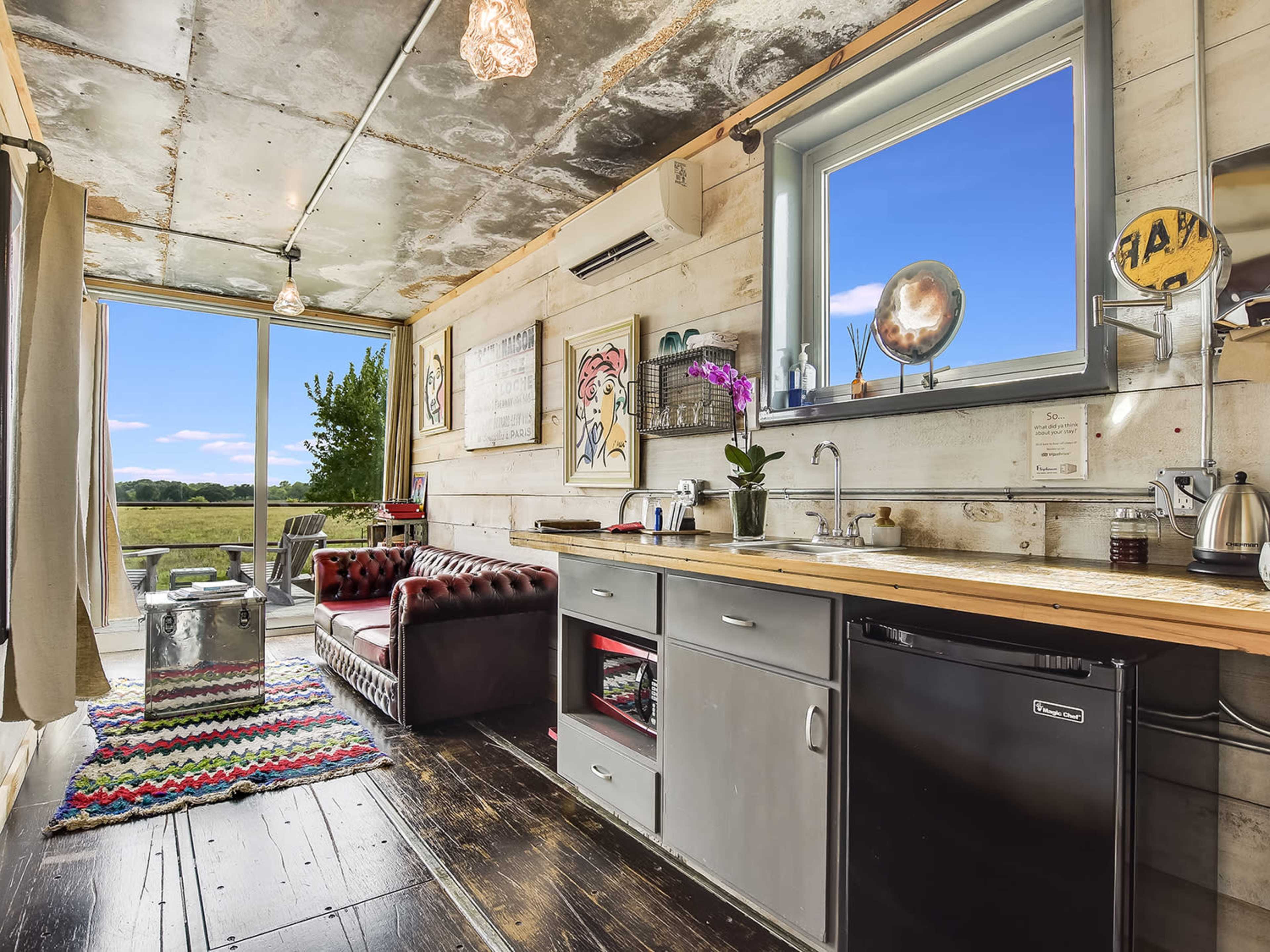 A small kitchen and living area with a sofa, wooden countertop, sink, and refrigerator, featuring a large window that overlooks a scenic landscape.