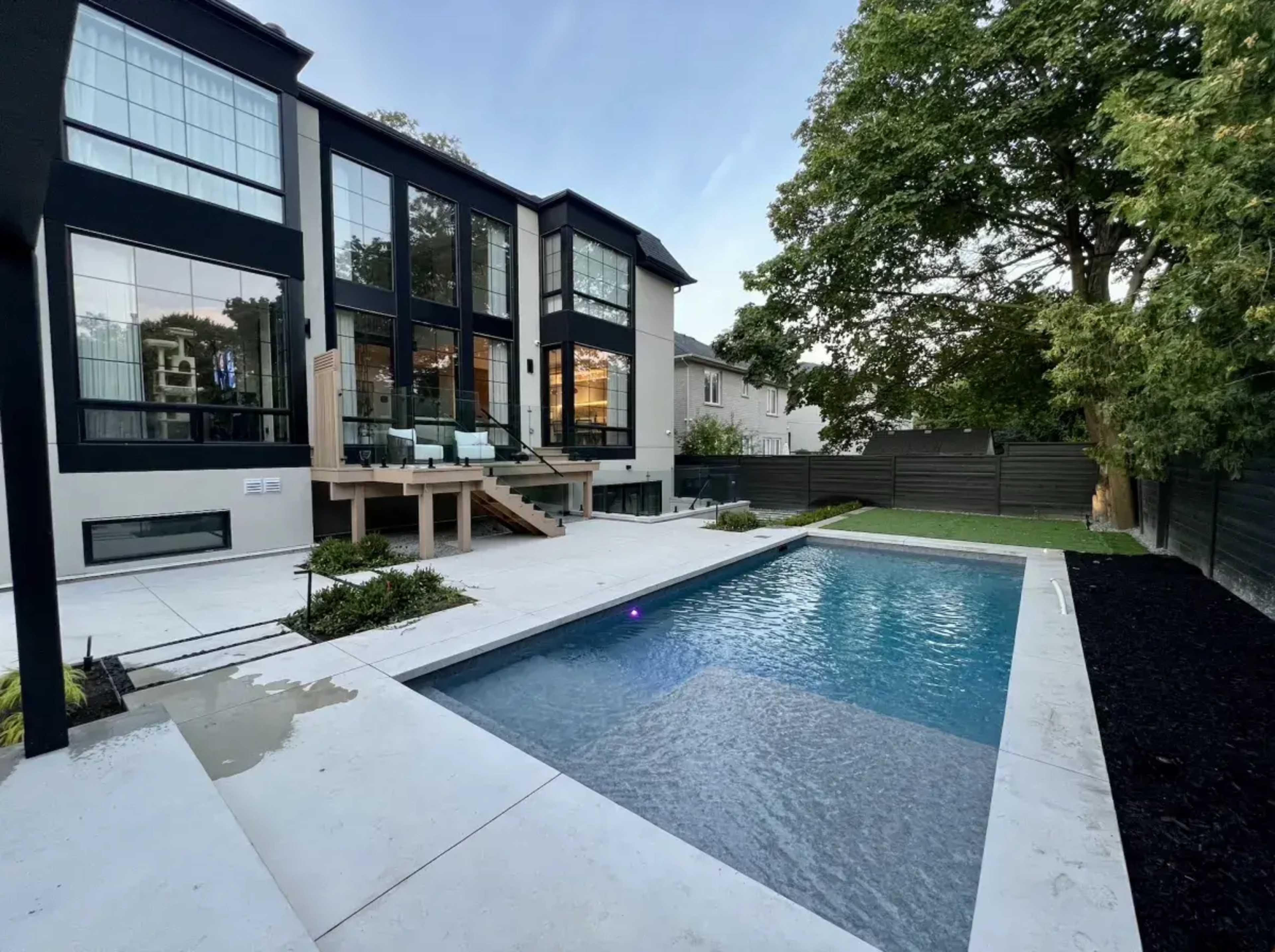 The image shows a modern house featuring large windows overlooking a rectangular swimming pool set within a landscaped yard.