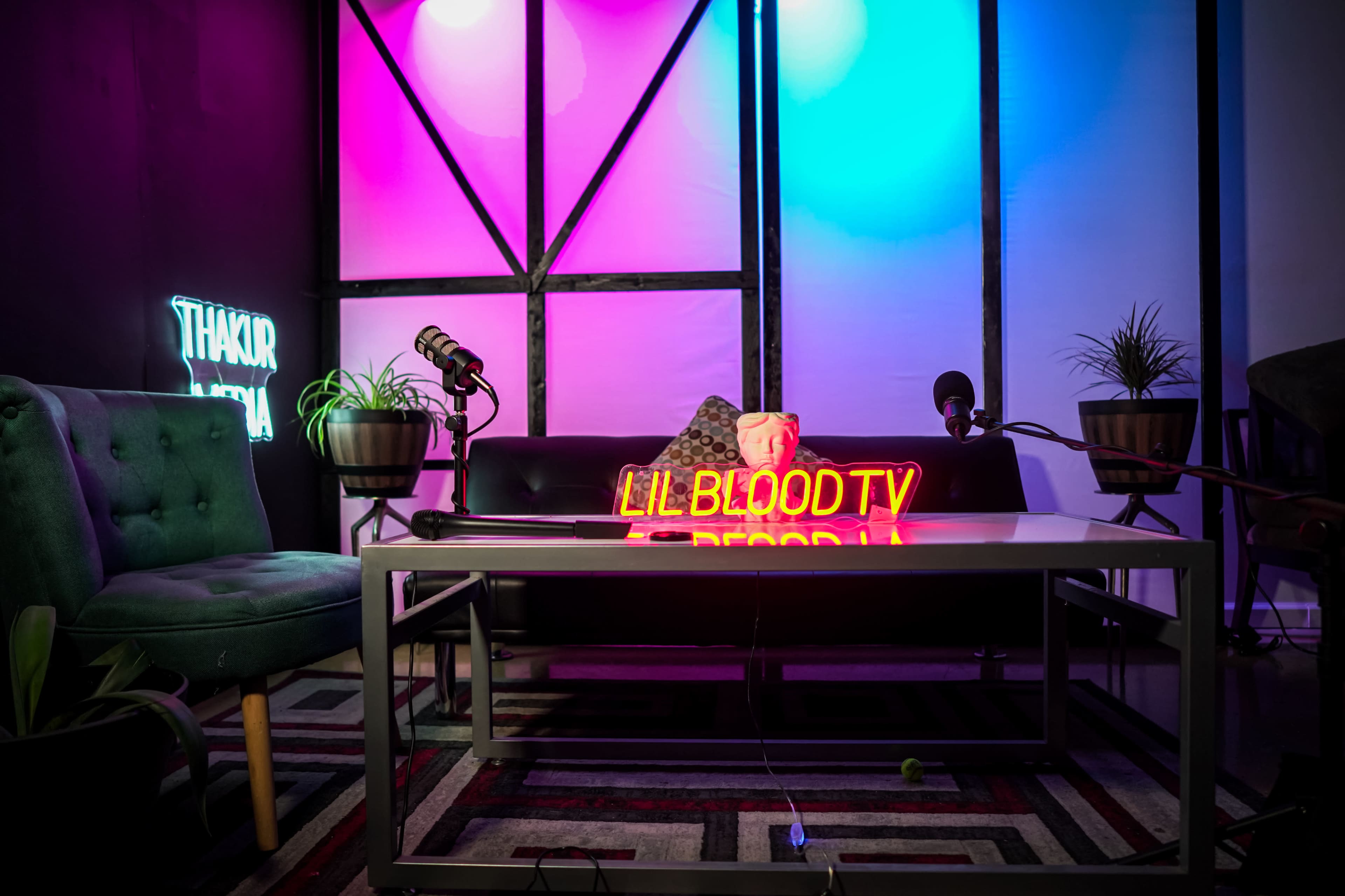 A stylized podcast set featuring a black sofa, a glass coffee table with a neon sign reading "LILBLOODTV," and several microphones arranged around the space.