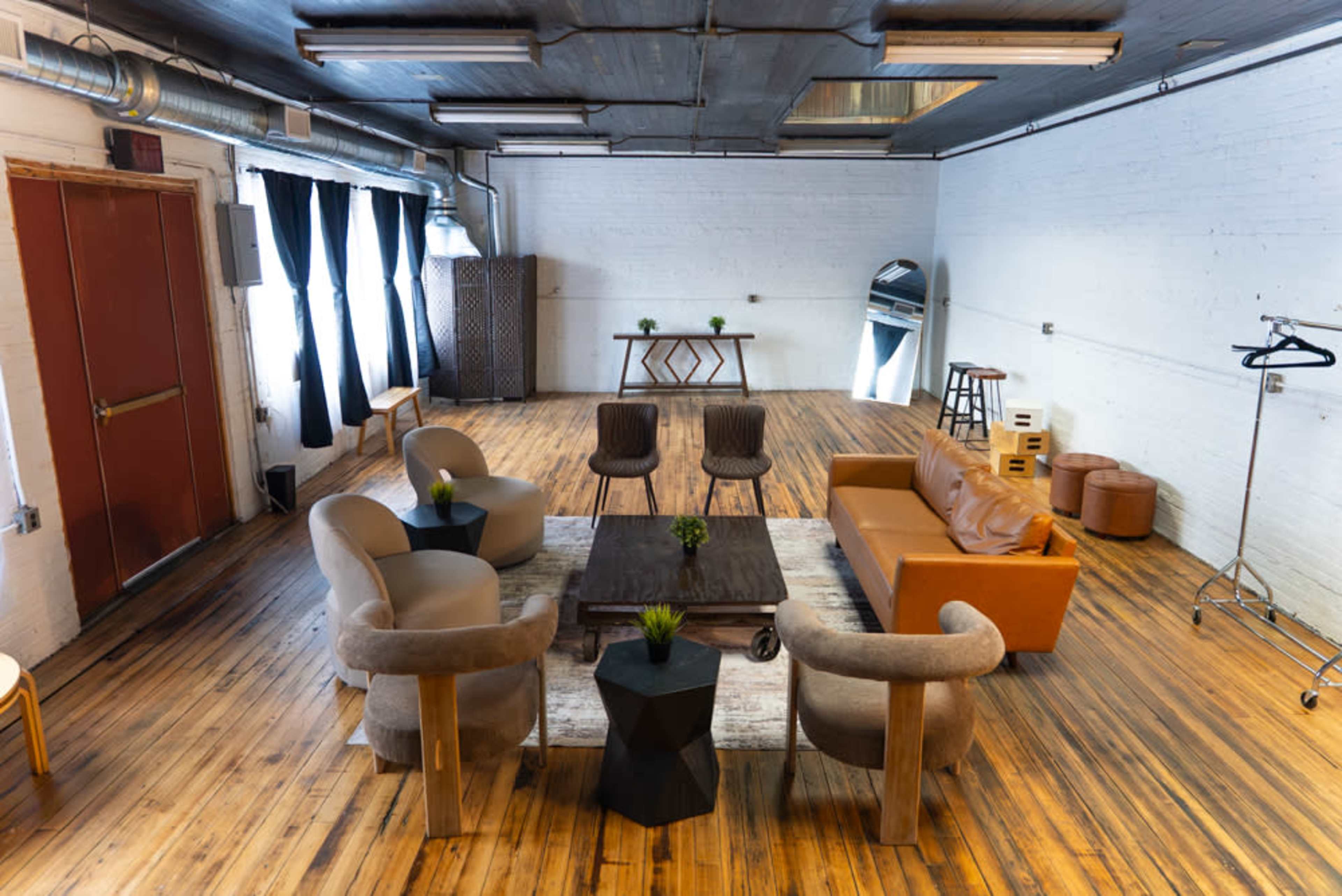 The image shows an open, modern interior space with wooden floors, a mix of seating arrangements, and decorative plants on a central table.