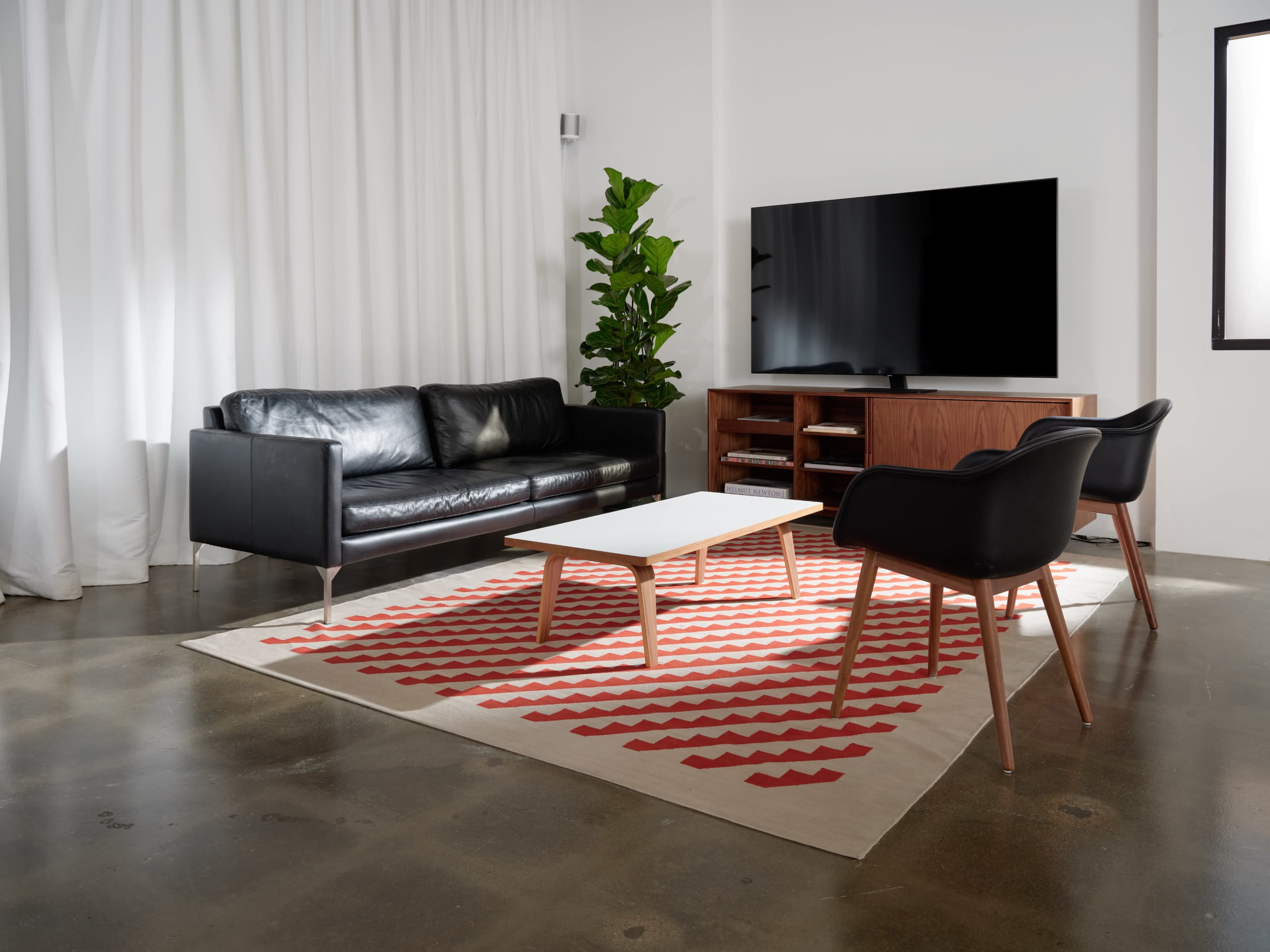 A modern living room features a black leather sofa, a white coffee table, and two black chairs on a patterned rug, with a television and a potted plant nearby.