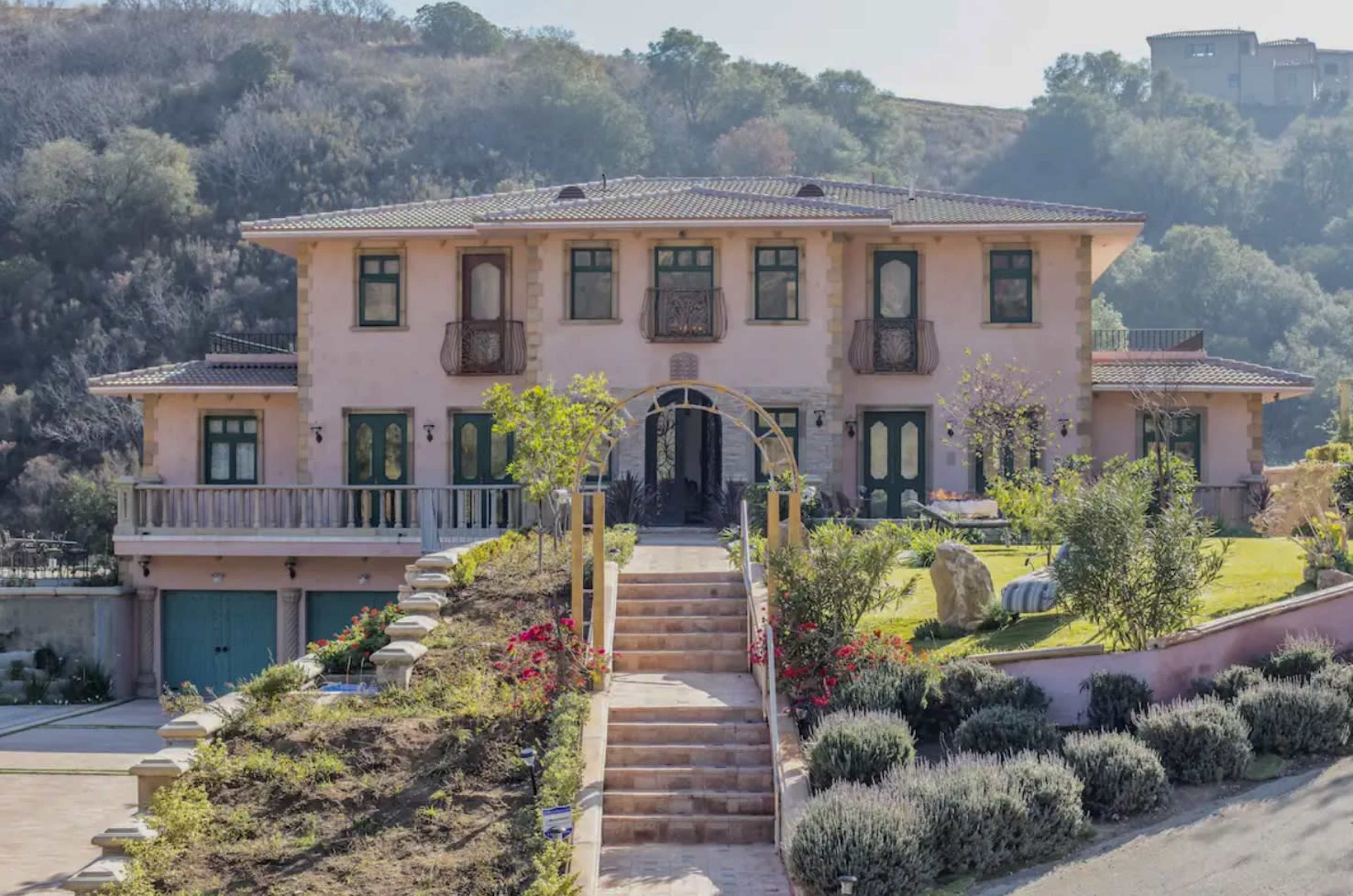 A large two-story house with a pink exterior, green shutters, and a landscaped walkway leads up to its entrance, surrounded by hills.