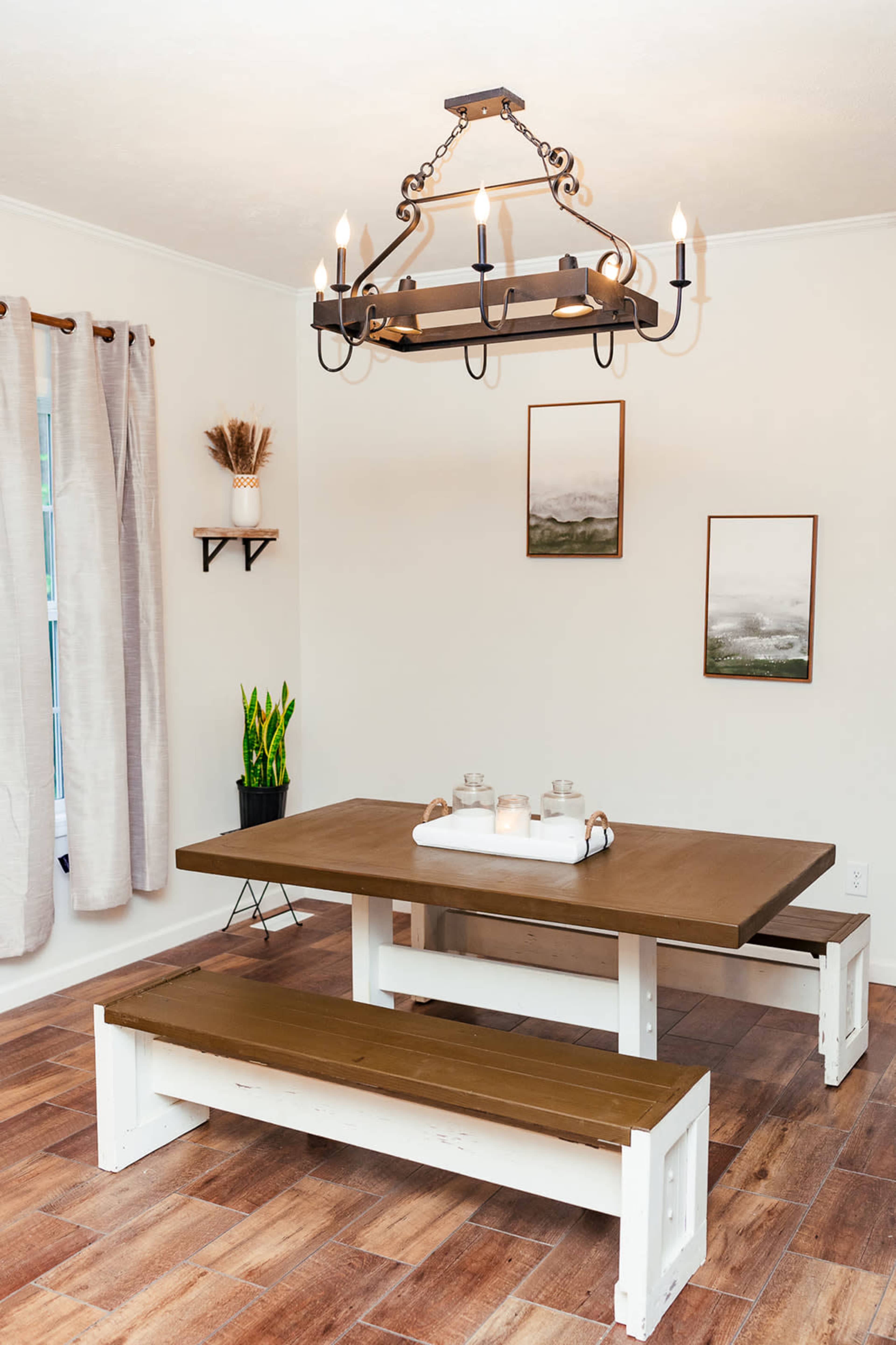 A wooden dining table with two benches is situated beneath a chandelier in a well-lit room, accented by window treatments and framed artwork on the walls.