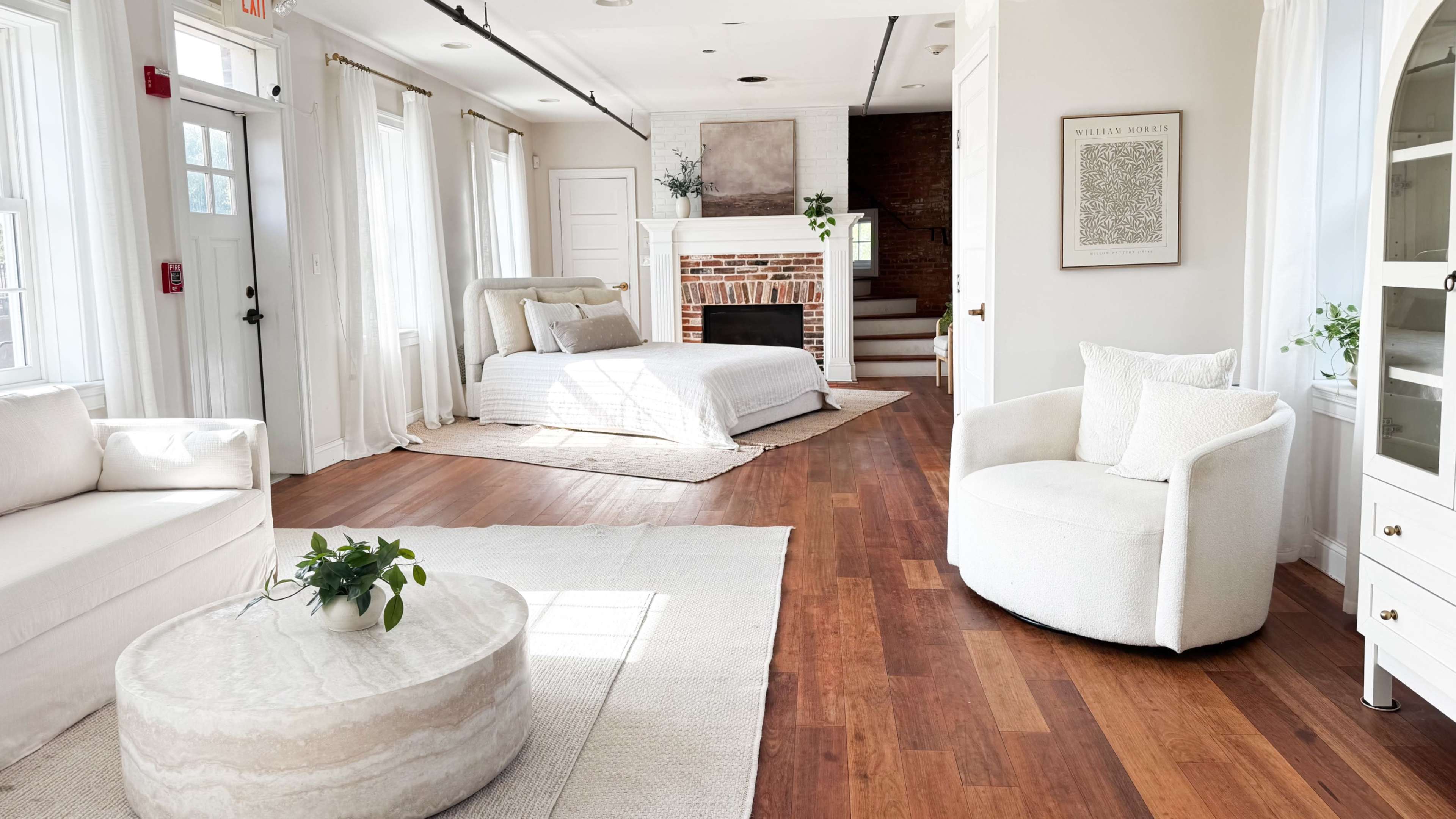 The image shows a bright, minimalist room featuring a bed, a circular table, and a cozy seating area with light-colored furniture.