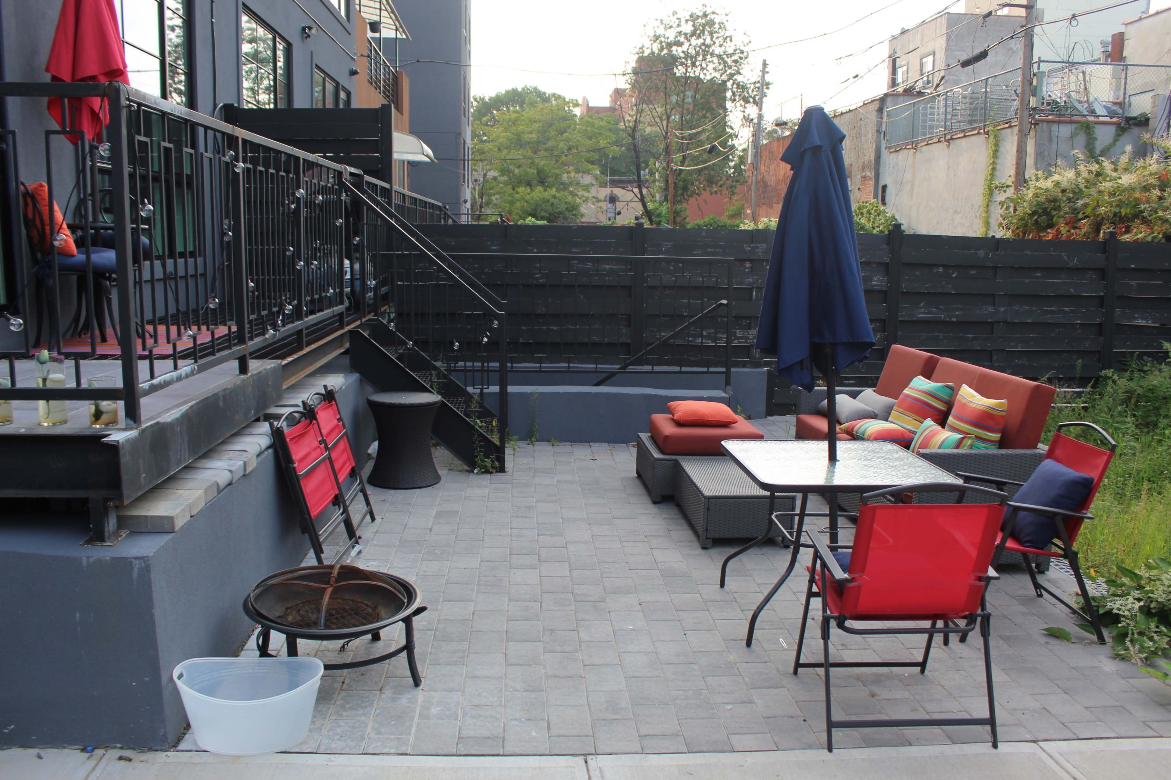 A patio area with a seating arrangement, an umbrella table, a fire pit, and red chairs alongside a black fenced wall.