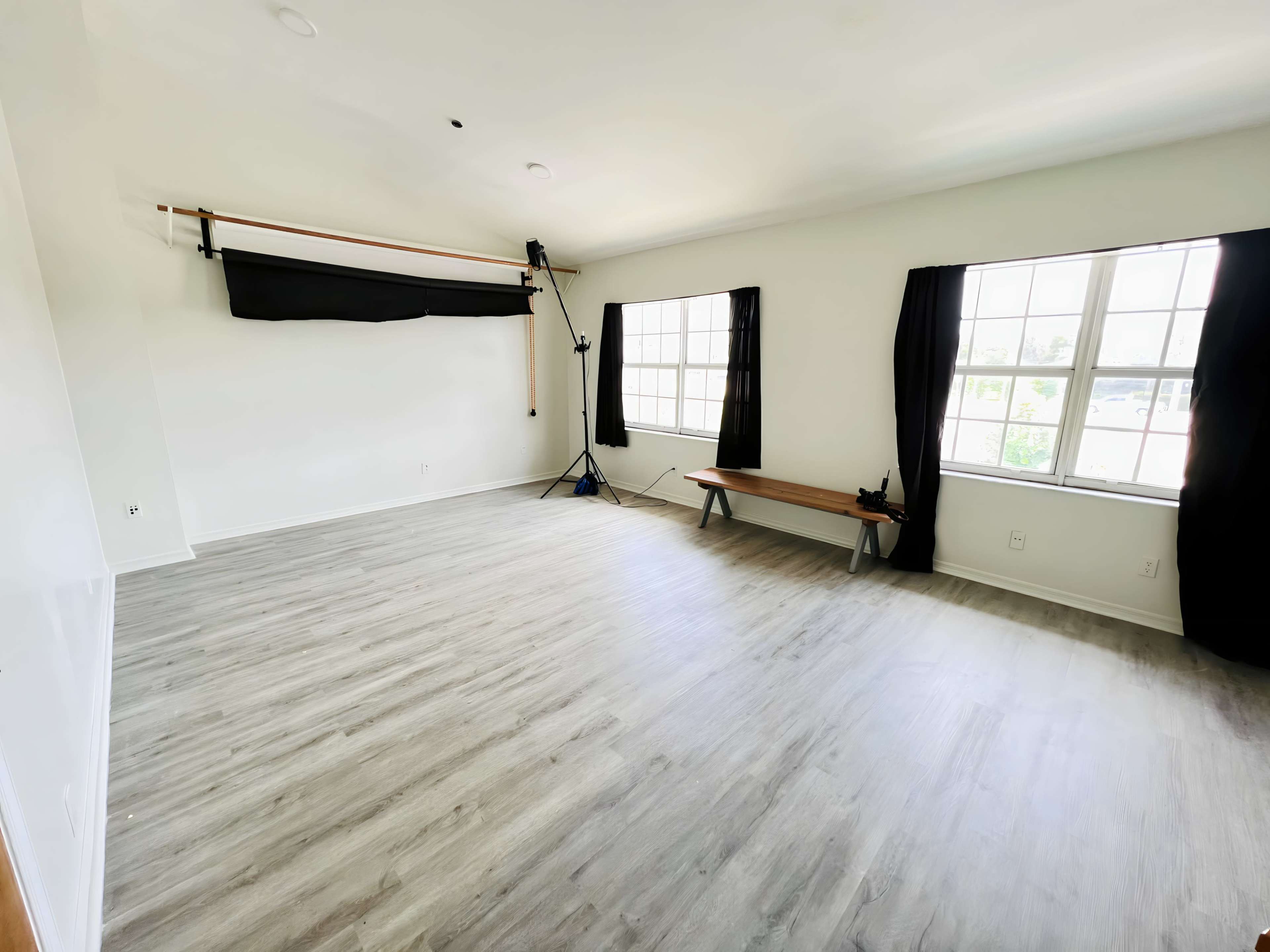 The image shows a vacant room with light-colored walls, a wooden floor, a bench against one wall, and large windows with black curtains.