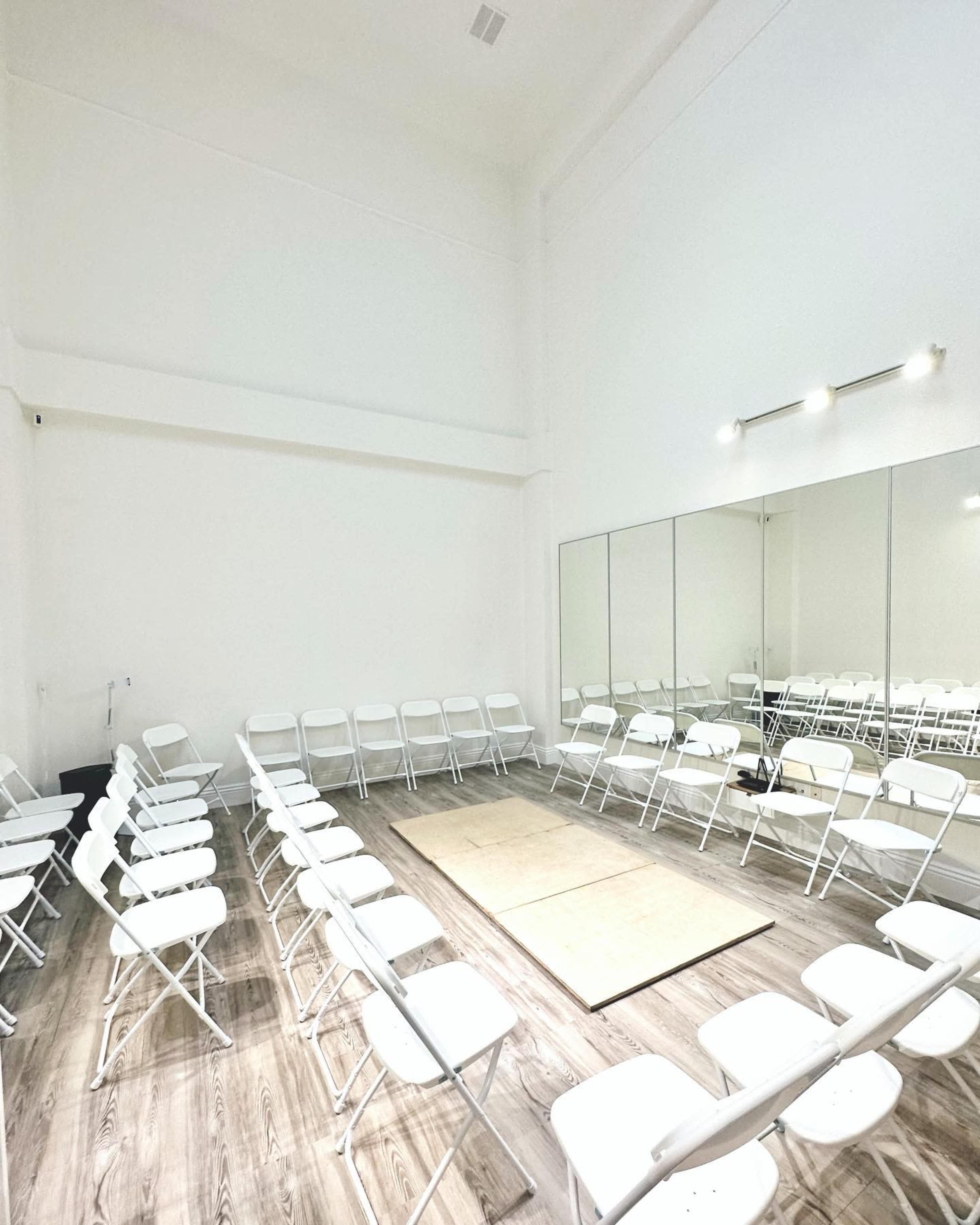 The image shows a minimalist room with white walls, a large mirror, and rows of white folding chairs arranged around a central space on a wooden floor.