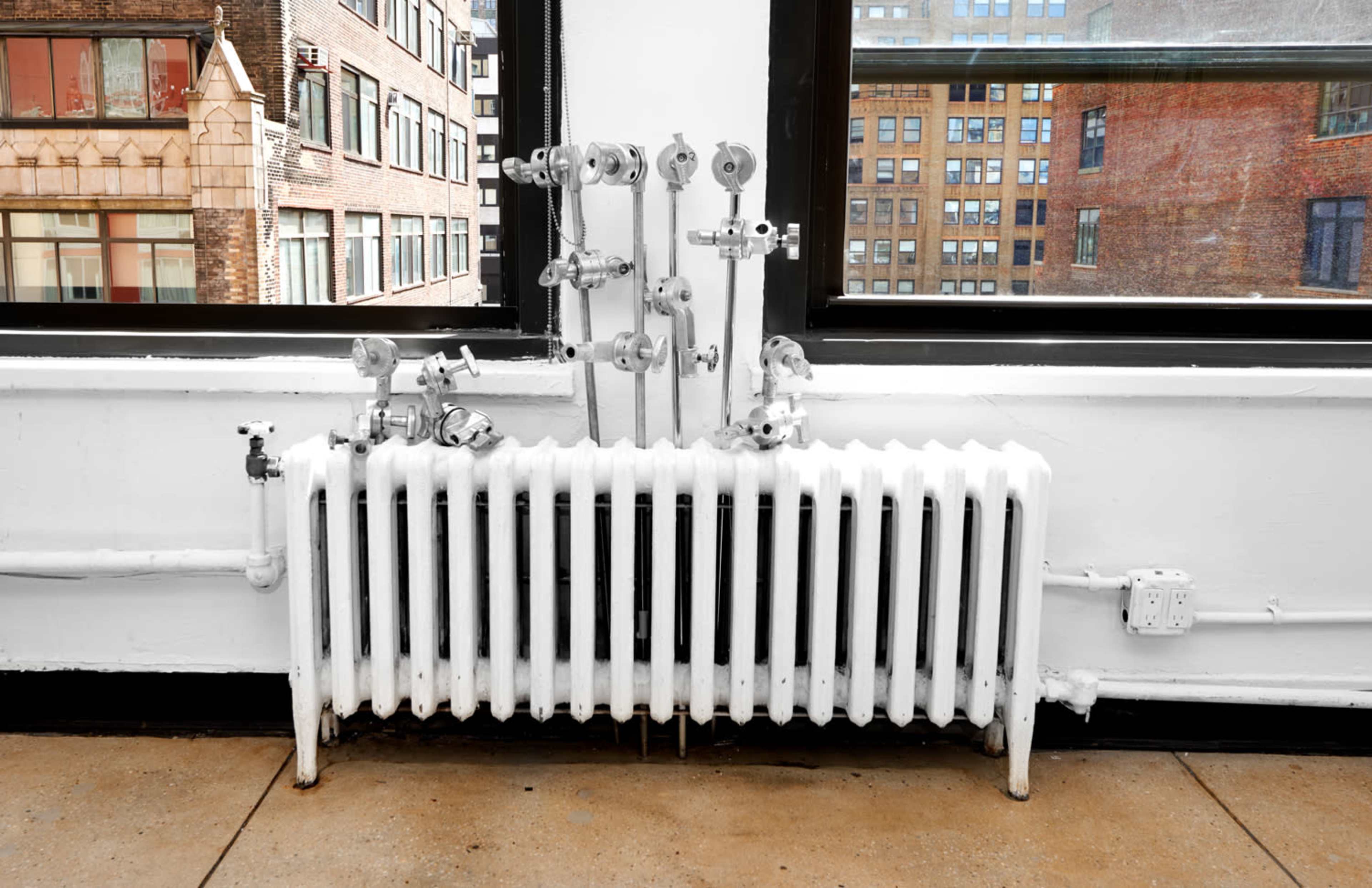 A white radiator with several valves is positioned below large windows in a modern building.