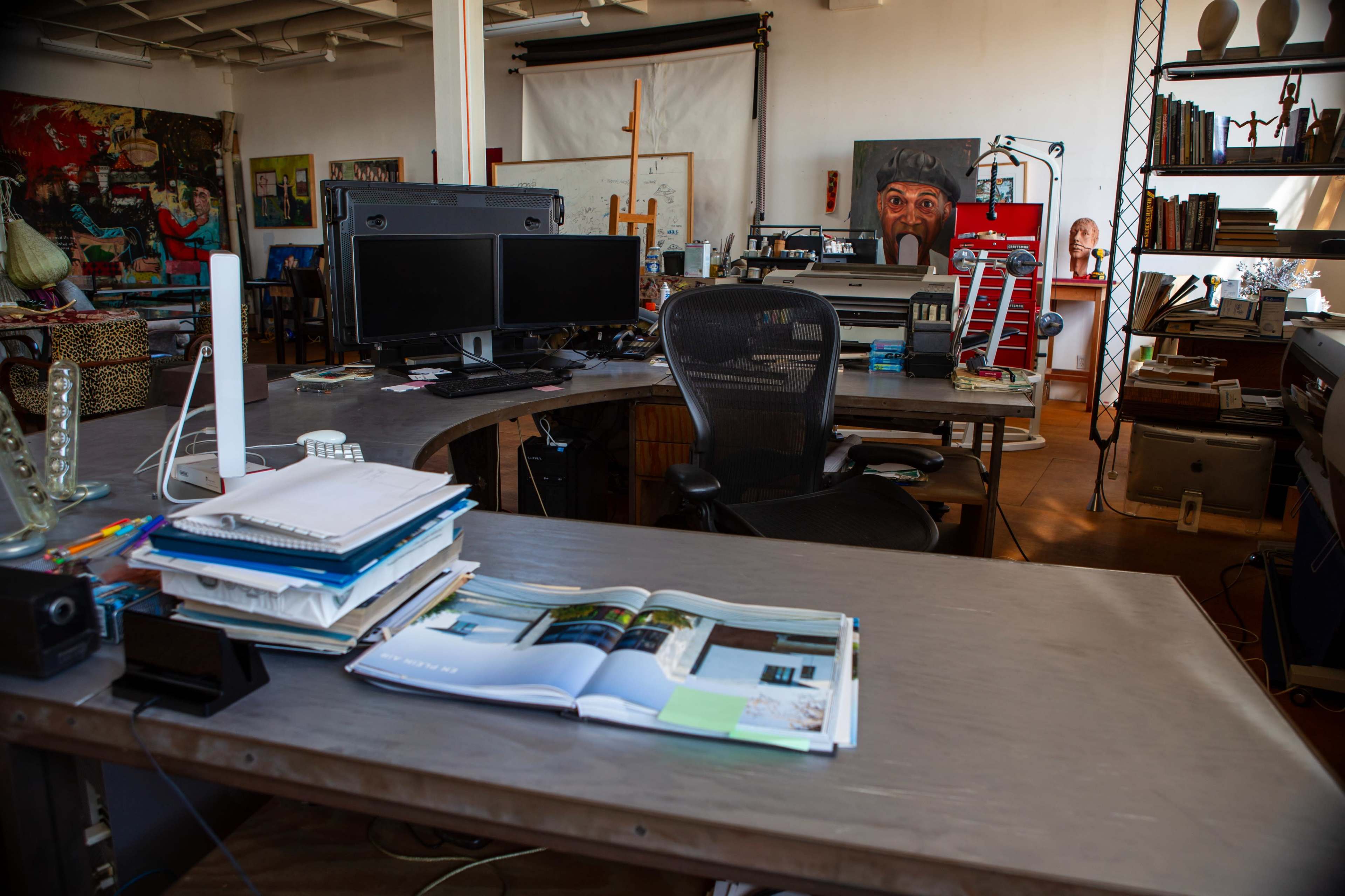 The image shows a spacious artist's studio with multiple workstations, two computer monitors, scattered art supplies, and paintings on the walls.
