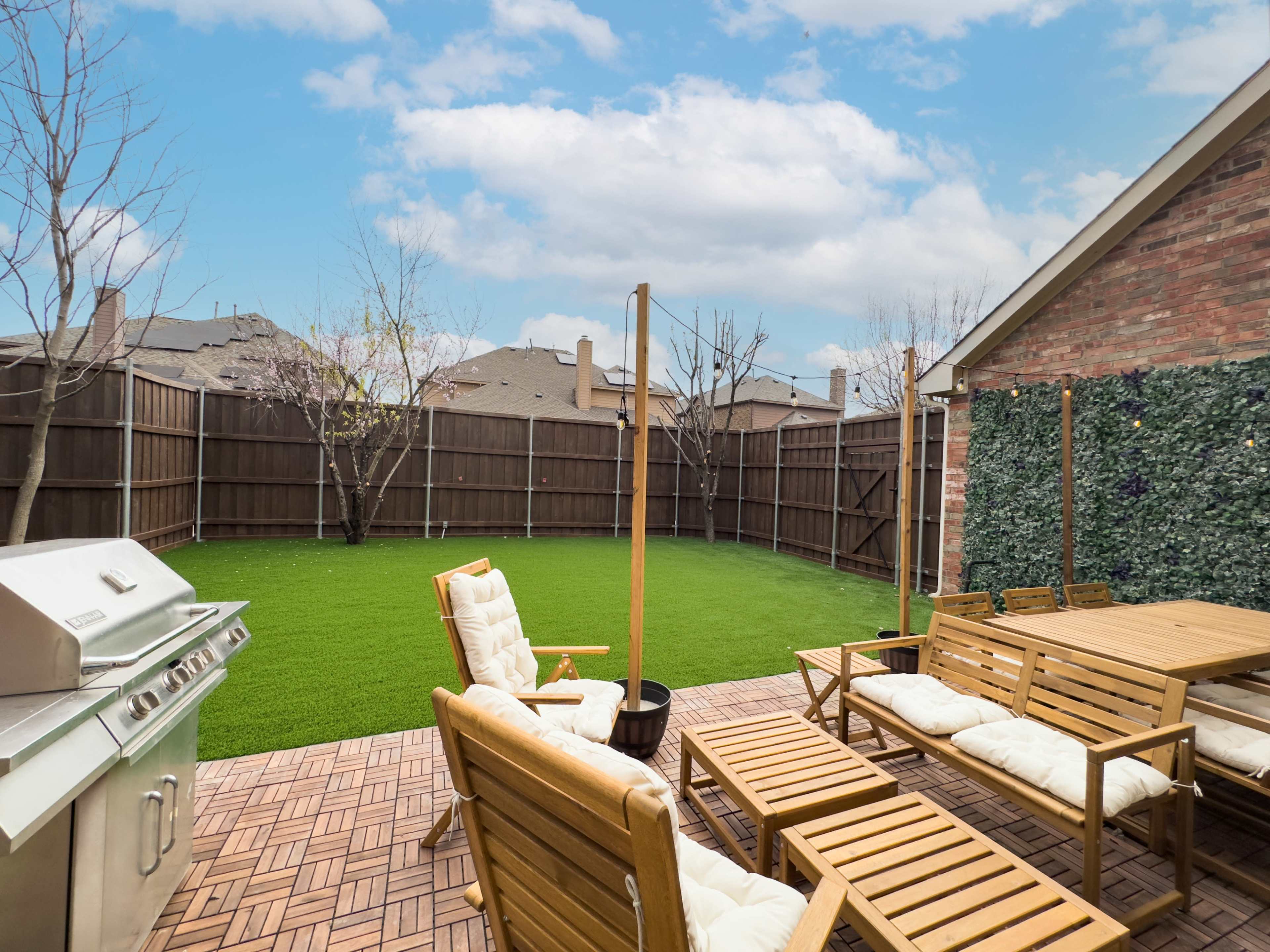 The image shows a backyard patio with wooden furniture and a barbecue grill, enclosed by a tall fence and featuring artificial grass.