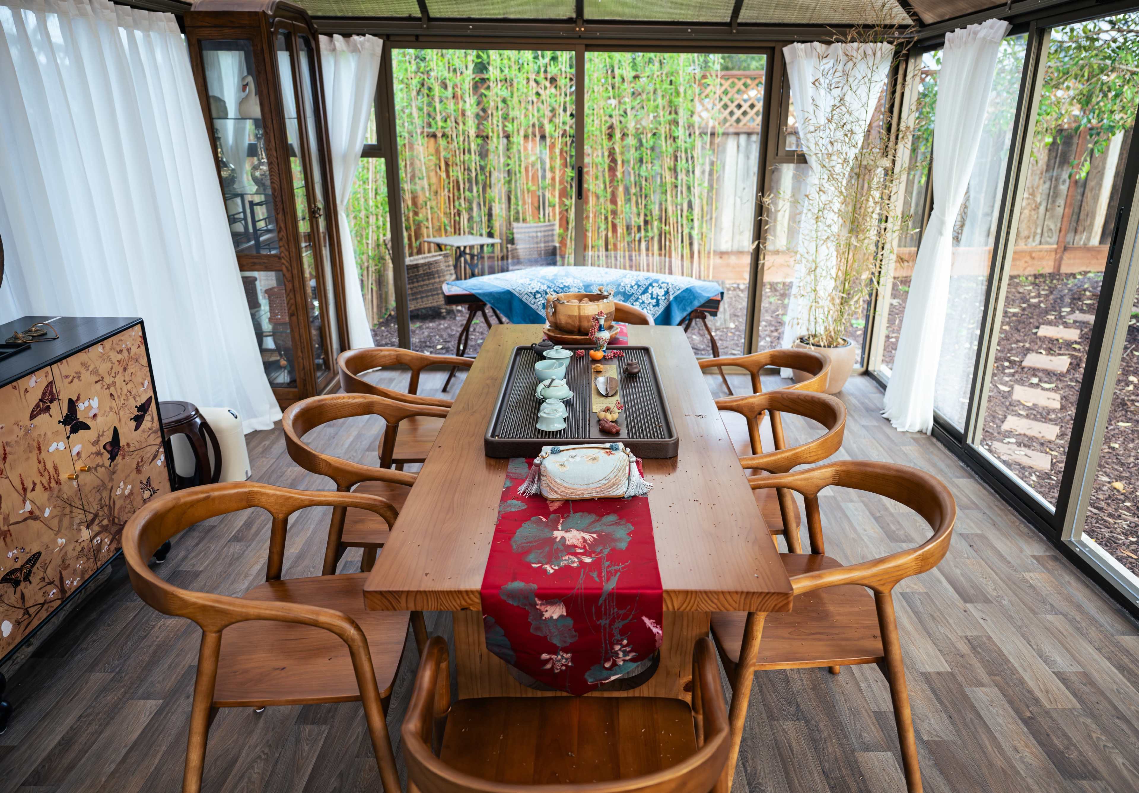 A wooden dining table is set in a glass-enclosed room, surrounded by chairs and featuring a decorative table runner, while sunlight filters through sheer curtains and bamboo plants are visible outside.