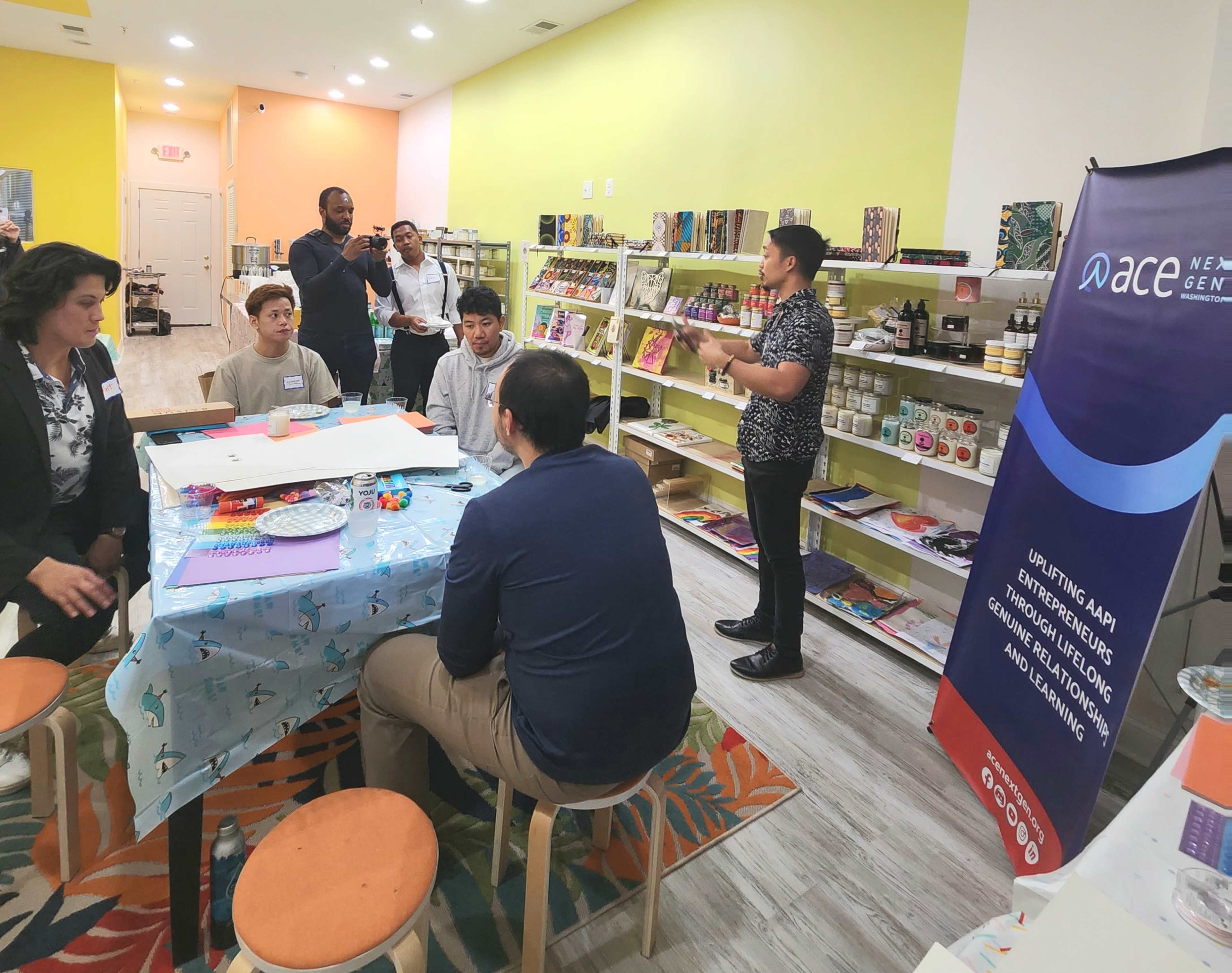 A group of people is seated around a table while a speaker stands in front of them in a brightly colored room filled with various products for sale.
