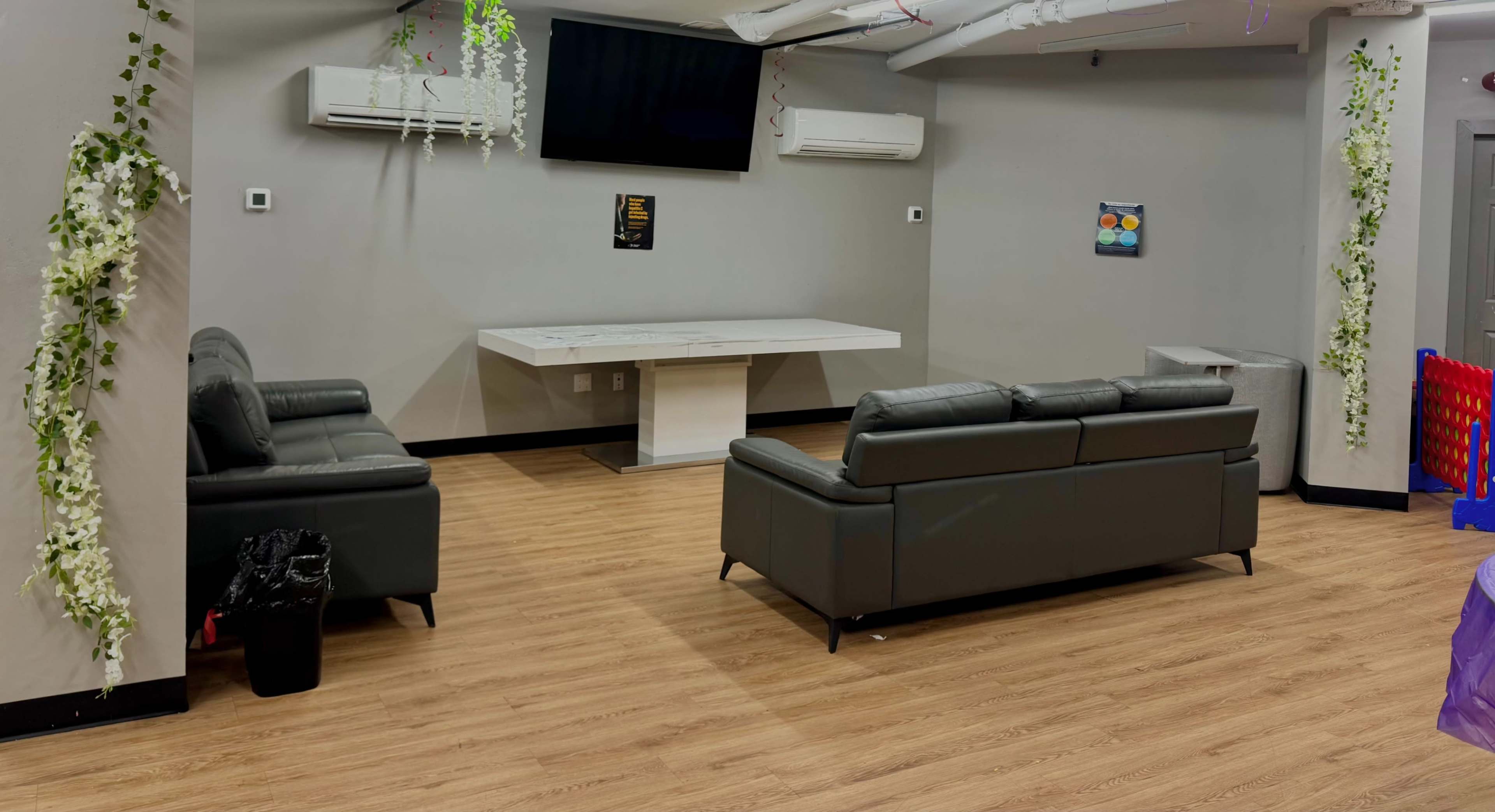 The image shows a modern lounge area with two black leather sofas, a light-colored table, and a wall-mounted TV in a room with wooden flooring and simple decor.
