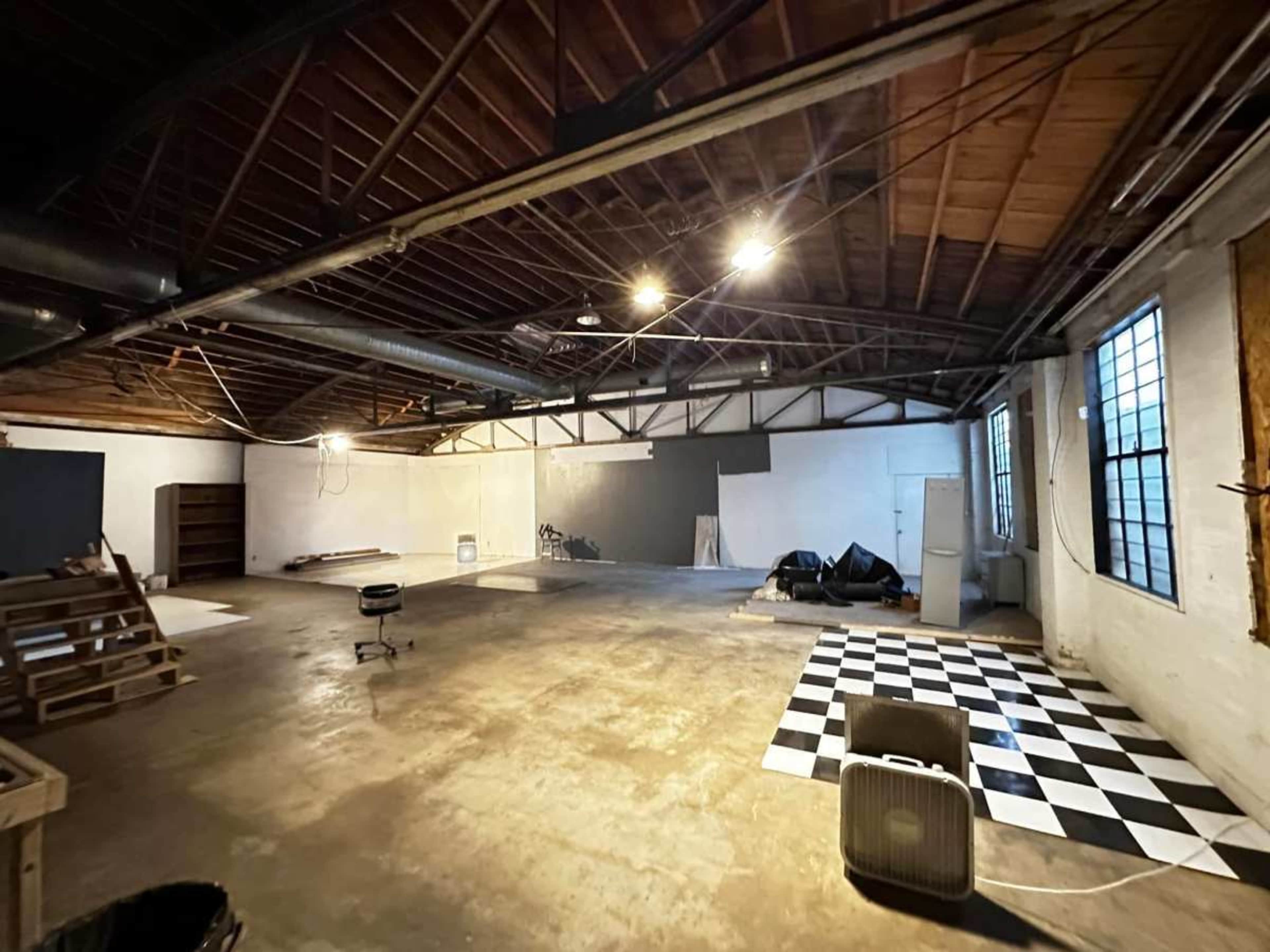 The image shows a spacious, unfinished interior of a warehouse or studio with wooden beams, scattered materials, and a black-and-white checkered pattern on the floor.
