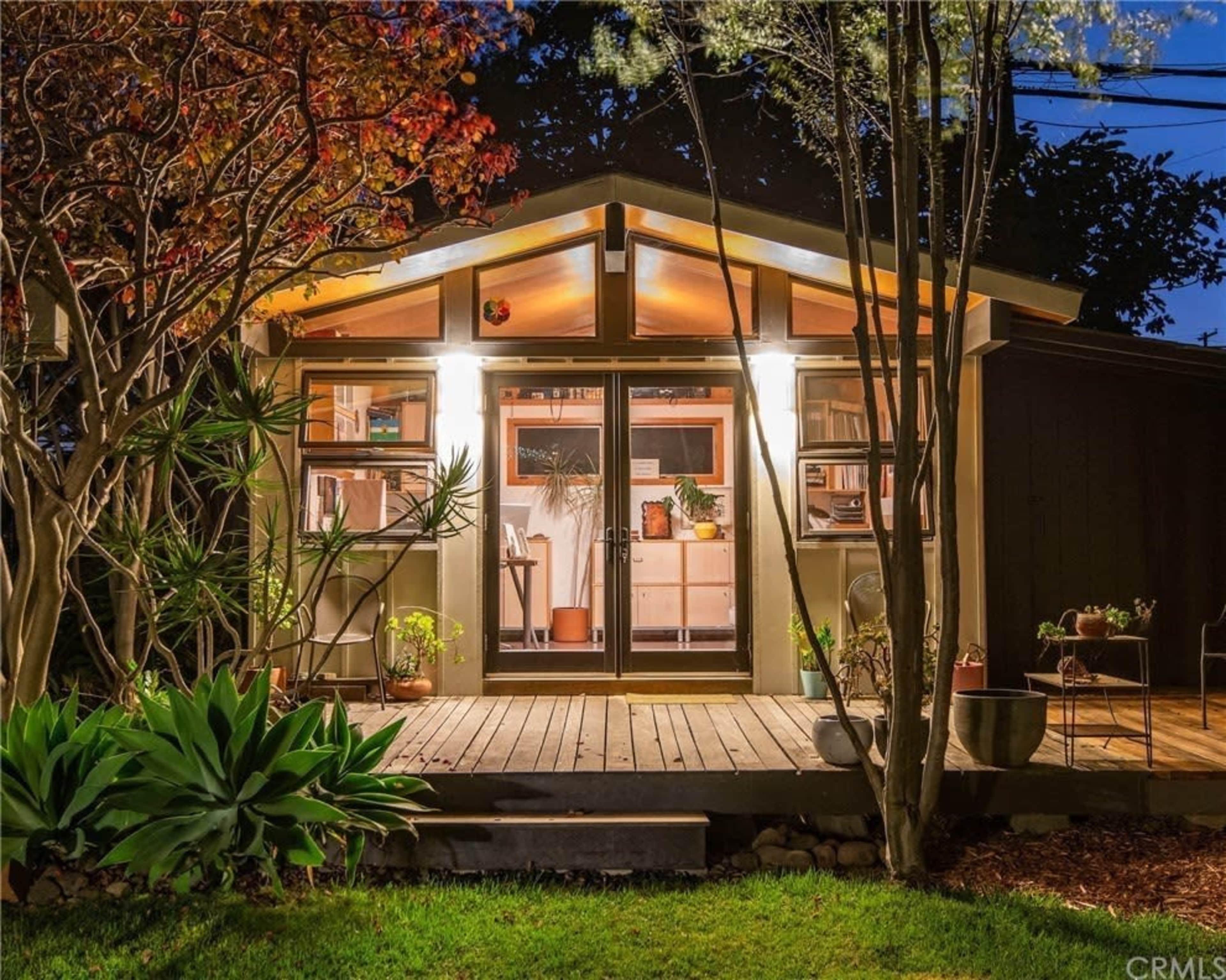 A cozy house with a wood-decked entryway features large glass doors and is surrounded by lush greenery and plants.