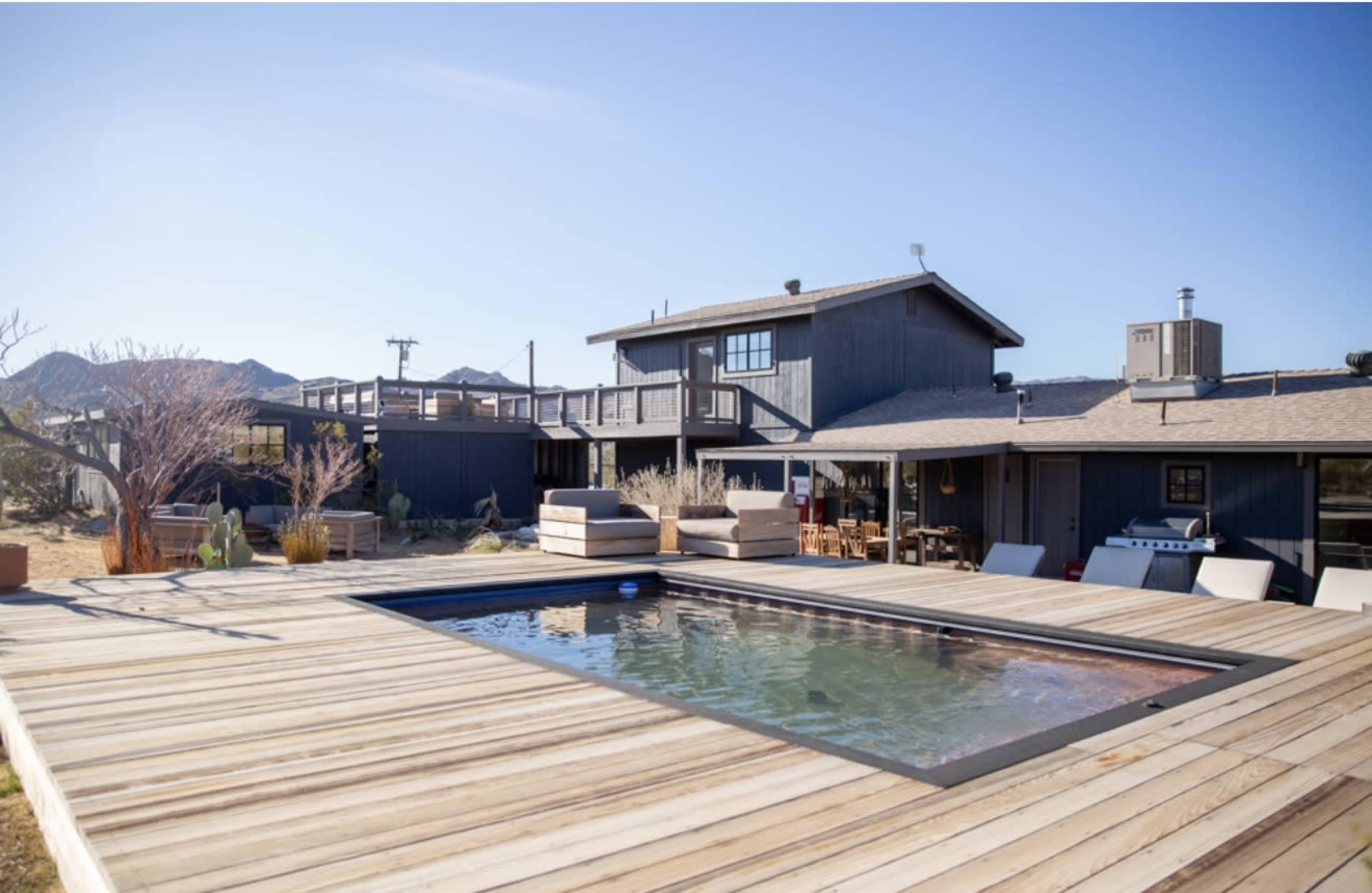 The image shows a modern house with a wooden deck and a rectangular swimming pool surrounded by lounge chairs in a desert landscape.