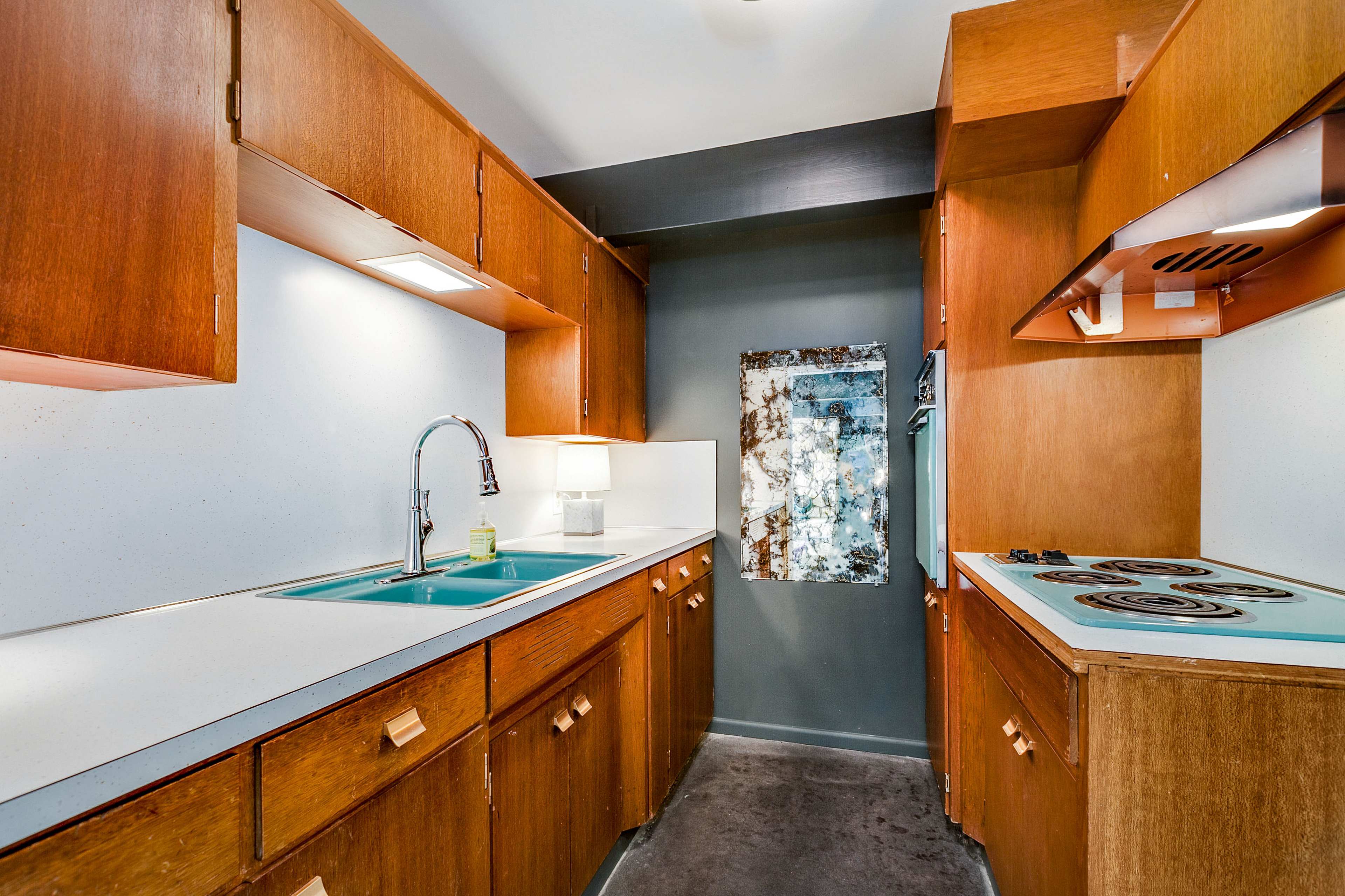 A narrow kitchen with wooden cabinets, a teal sink, and a stovetop against a gray wall.
